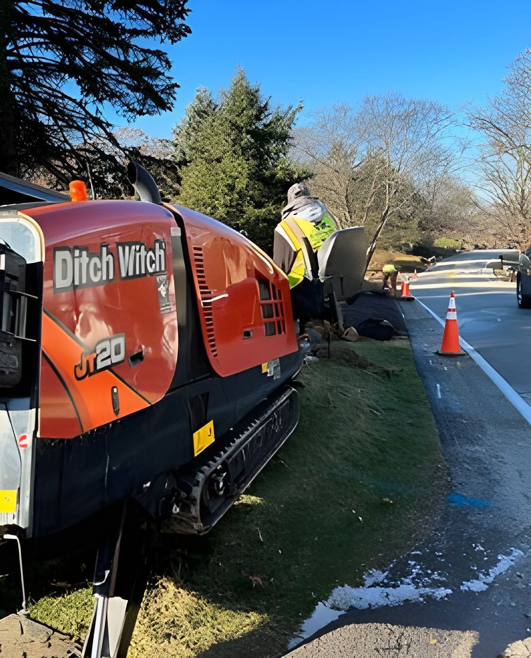 Orange “Ditch Witch” machine beside a road, with a worker and traffic cones near a roadside ditch.