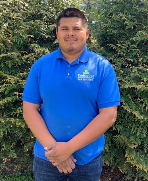 Man standing outdoors in a blue polo shirt in front of green bushes, hands clasped and smiling.