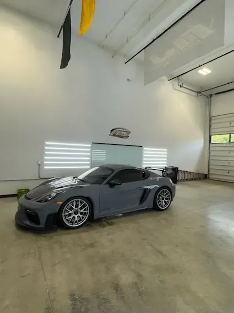A gray Porsche 718 Cayman GT4 RS parked in a bright, modern garage with white walls and a concrete floor.