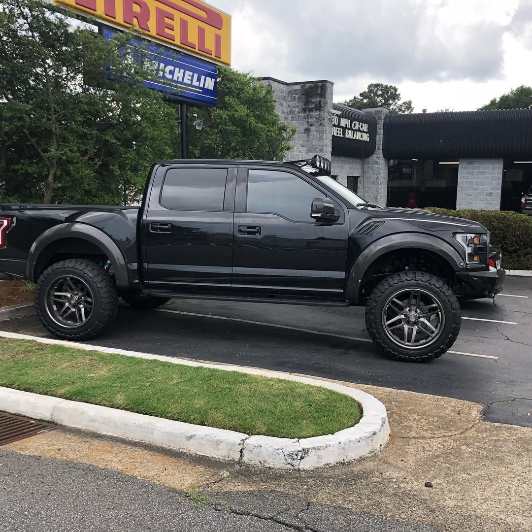 A black Ford F-150 Raptor pickup truck with a lifted suspension and custom wheels parked at an auto repair shop.
