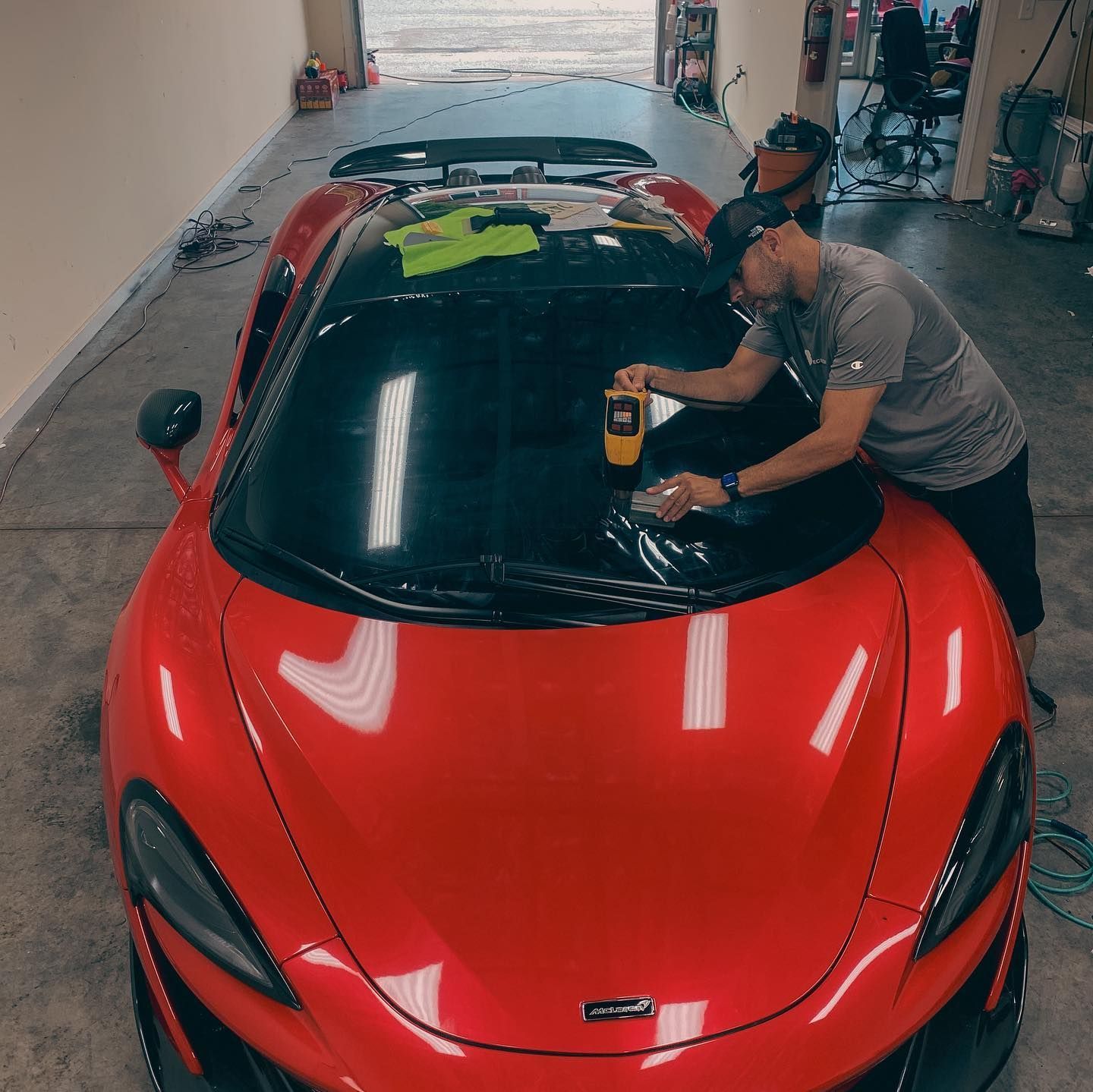 A person in a gray shirt uses a heat gun to apply window tint to the windshield of a bright red McLaren in a garage.