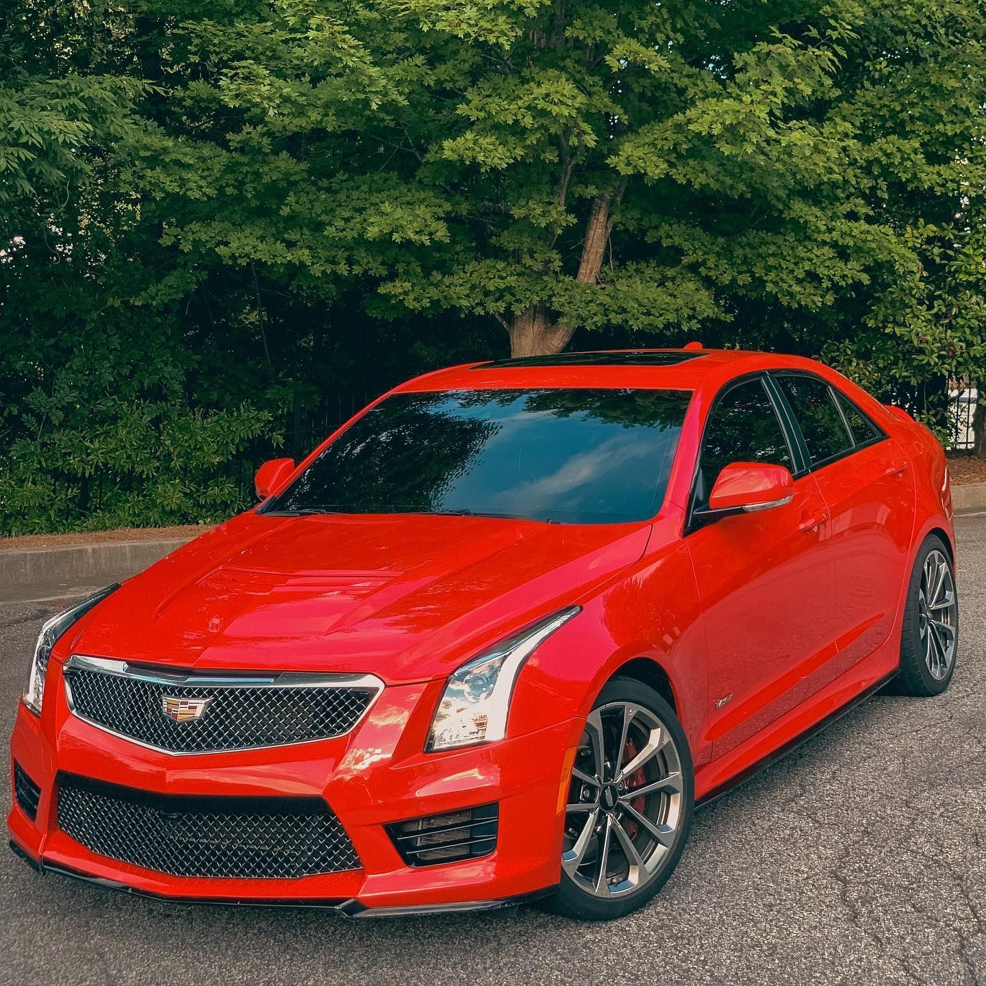 A bright red Cadillac ATS-V parked on an asphalt surface in front of green leafy trees.
