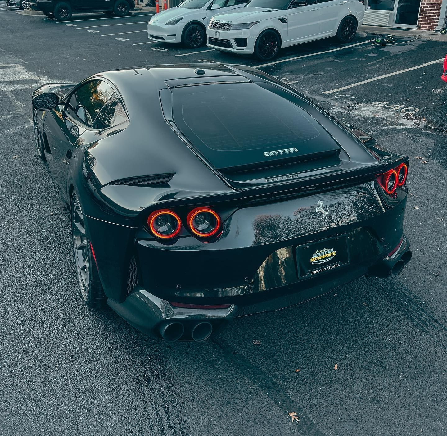 A black Ferrari sports car parked in a parking lot, with several other vehicles behind it near a building.