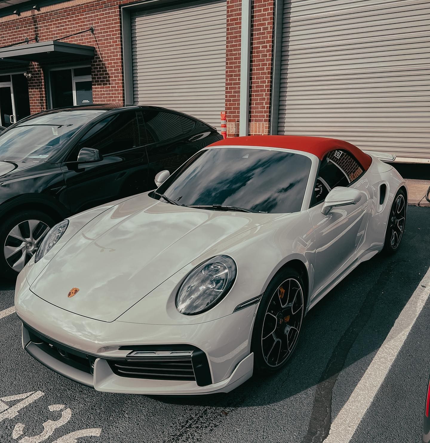 A white Porsche 911 convertible with a red top parked in a lot next to a black car in front of a brick building.