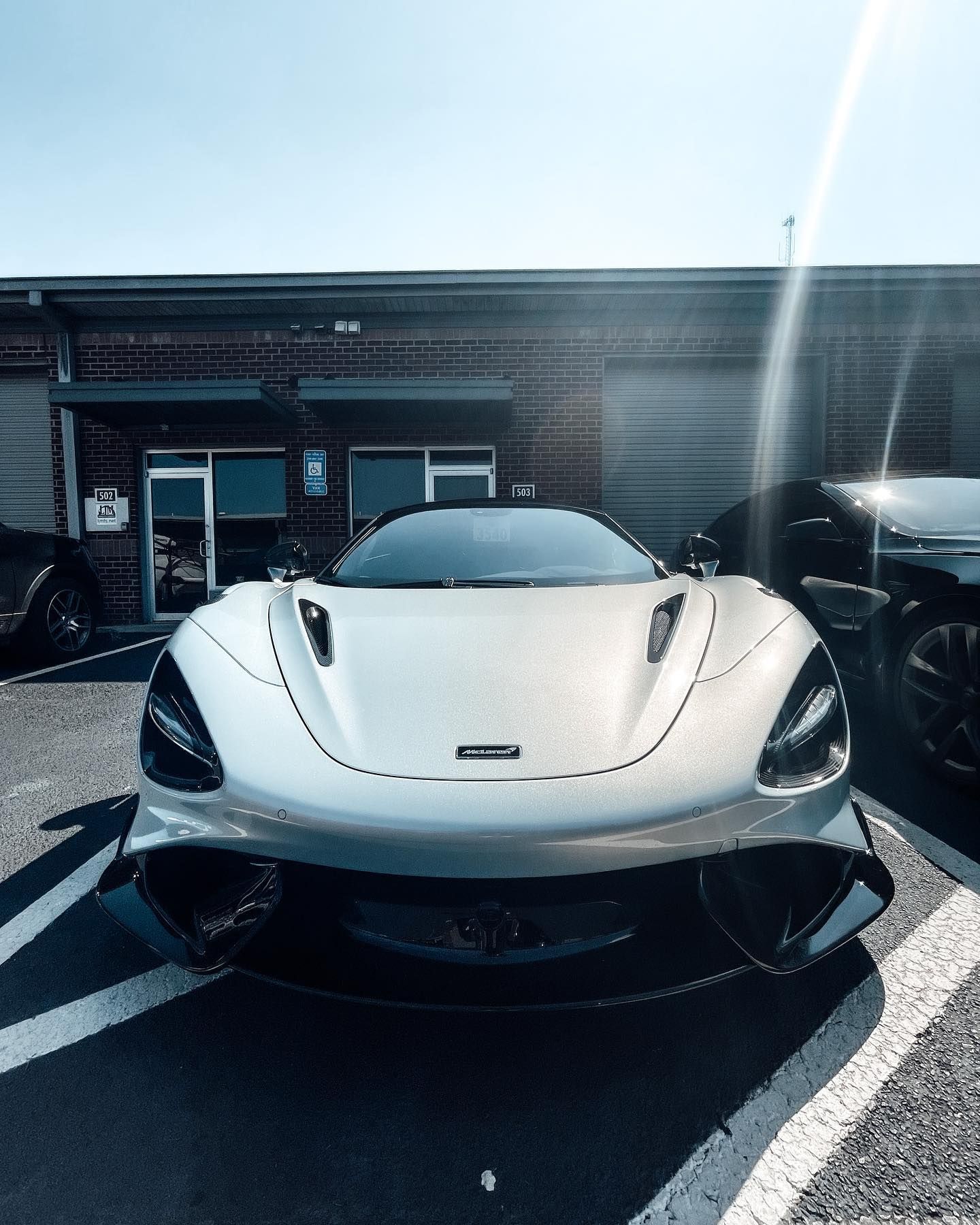 A silver McLaren sports car parked in an asphalt lot in front of a brick building on a sunny day.