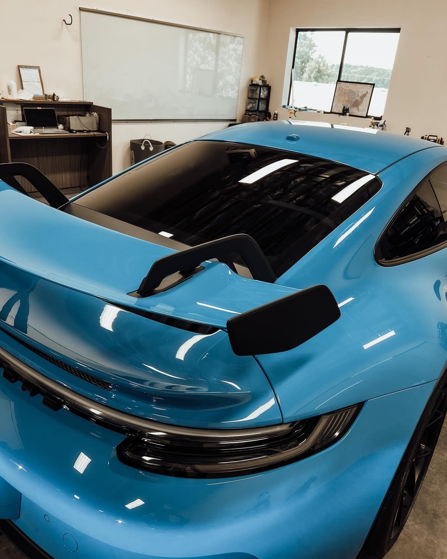 Rear view of a bright blue Porsche 911 GT3 with a prominent black rear wing, parked inside an office or garage.