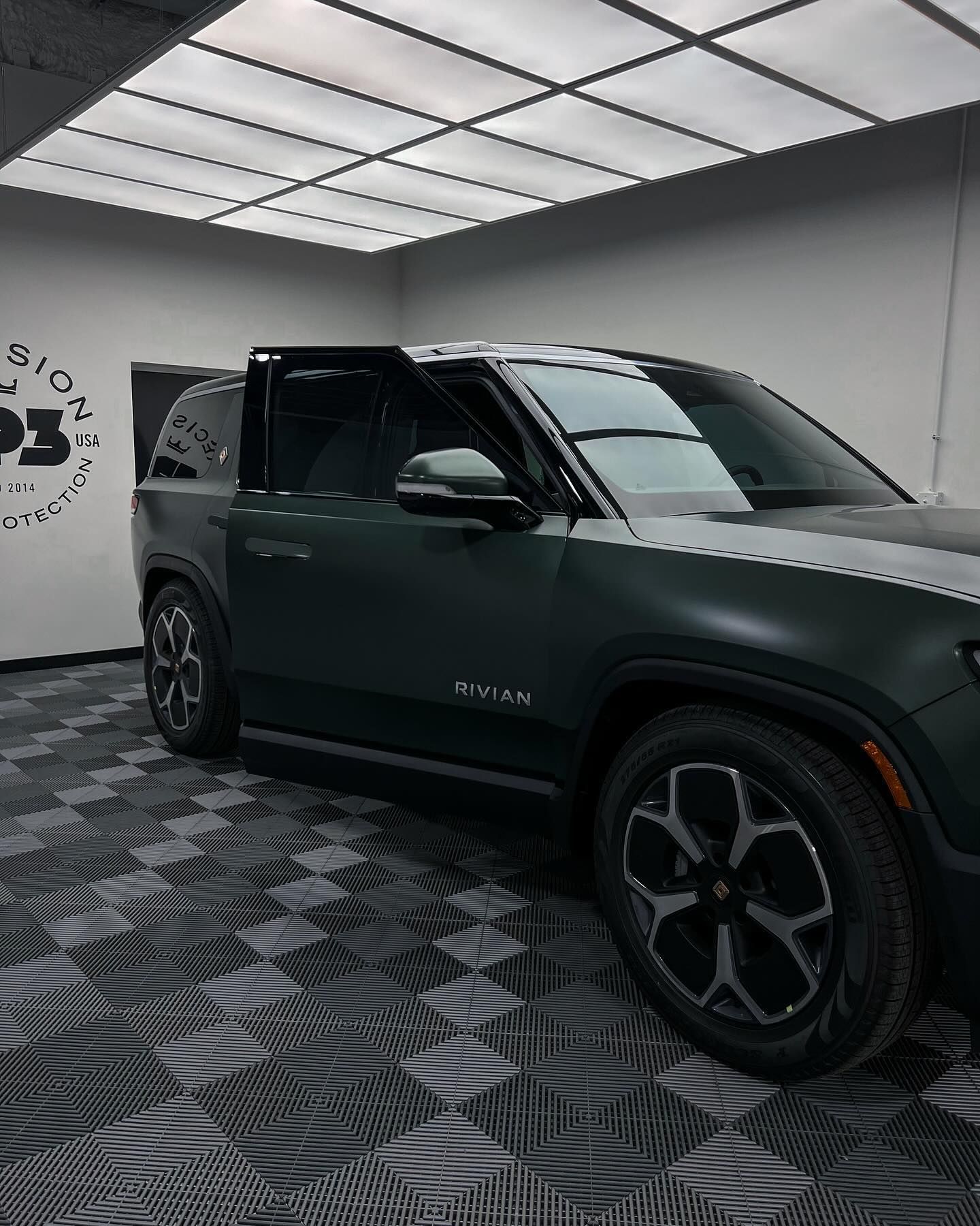 Matte dark green SUV parked in a bright studio with a checkered floor, side door slightly open.
