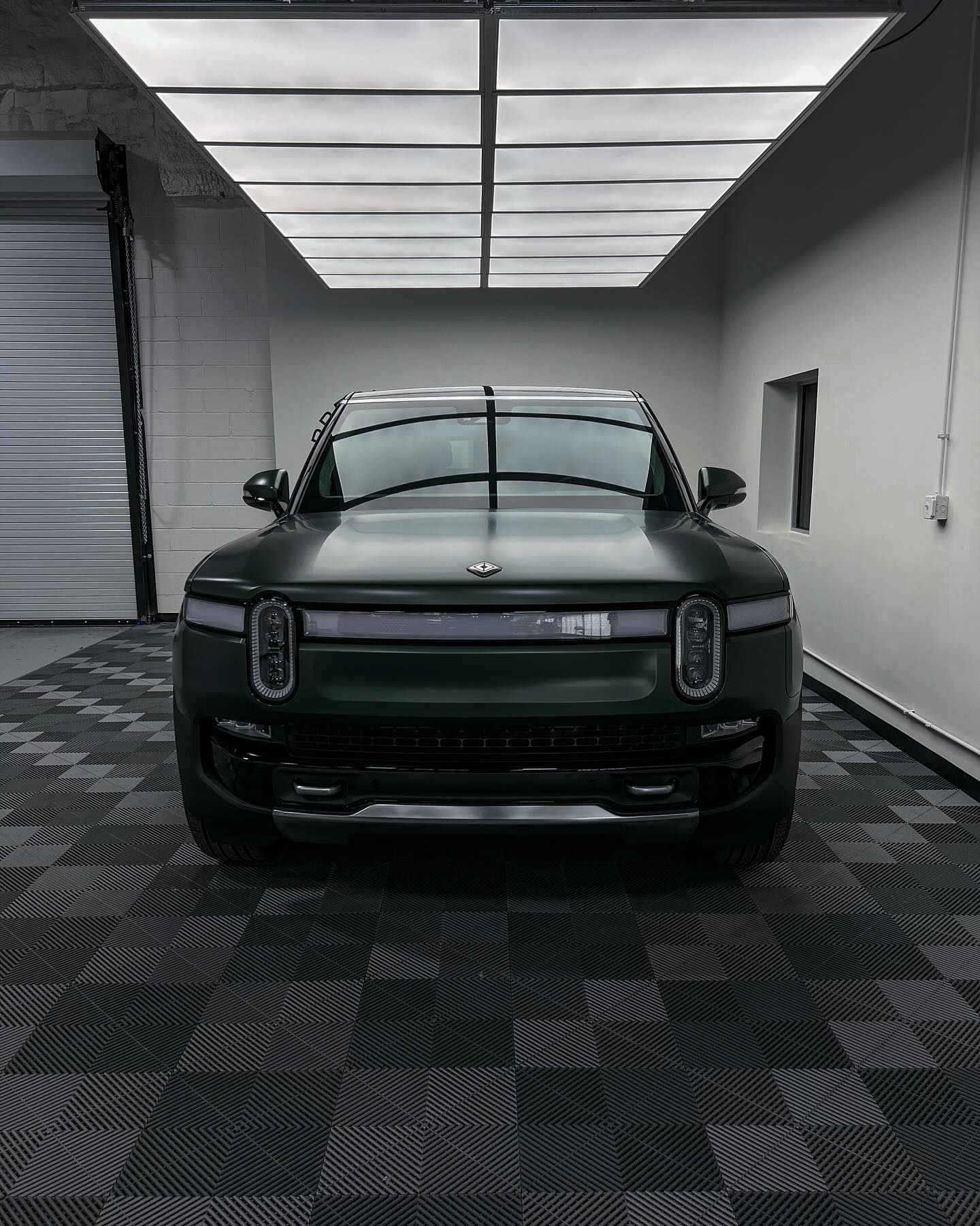 A dark green Rivian SUV parked in a modern, brightly lit garage with checkered flooring.
