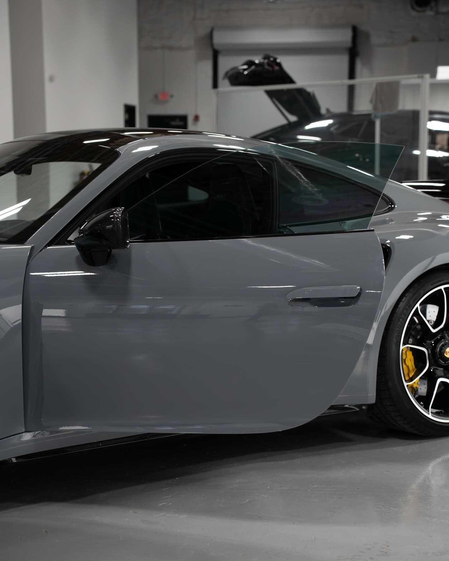 A gray sports car with its driver-side door open, parked in a bright garage with a partial view of another car behind.