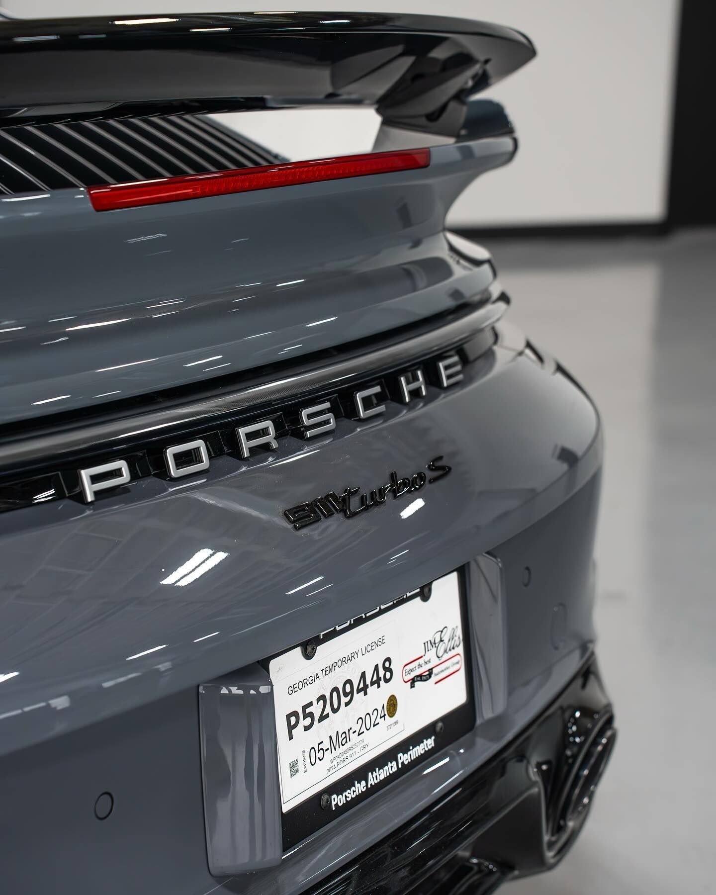 Rear view of a grey Porsche 911 Turbo S with a rear spoiler and temporary dealership tag in a garage.
