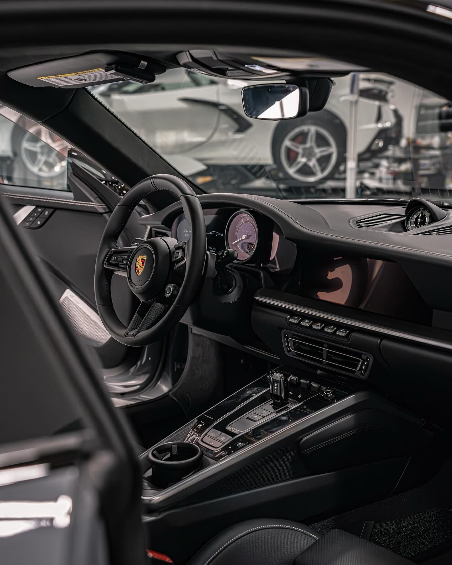 Interior of a dark-toned Porsche cockpit featuring a steering wheel with the brand logo and a carbon fiber center console.