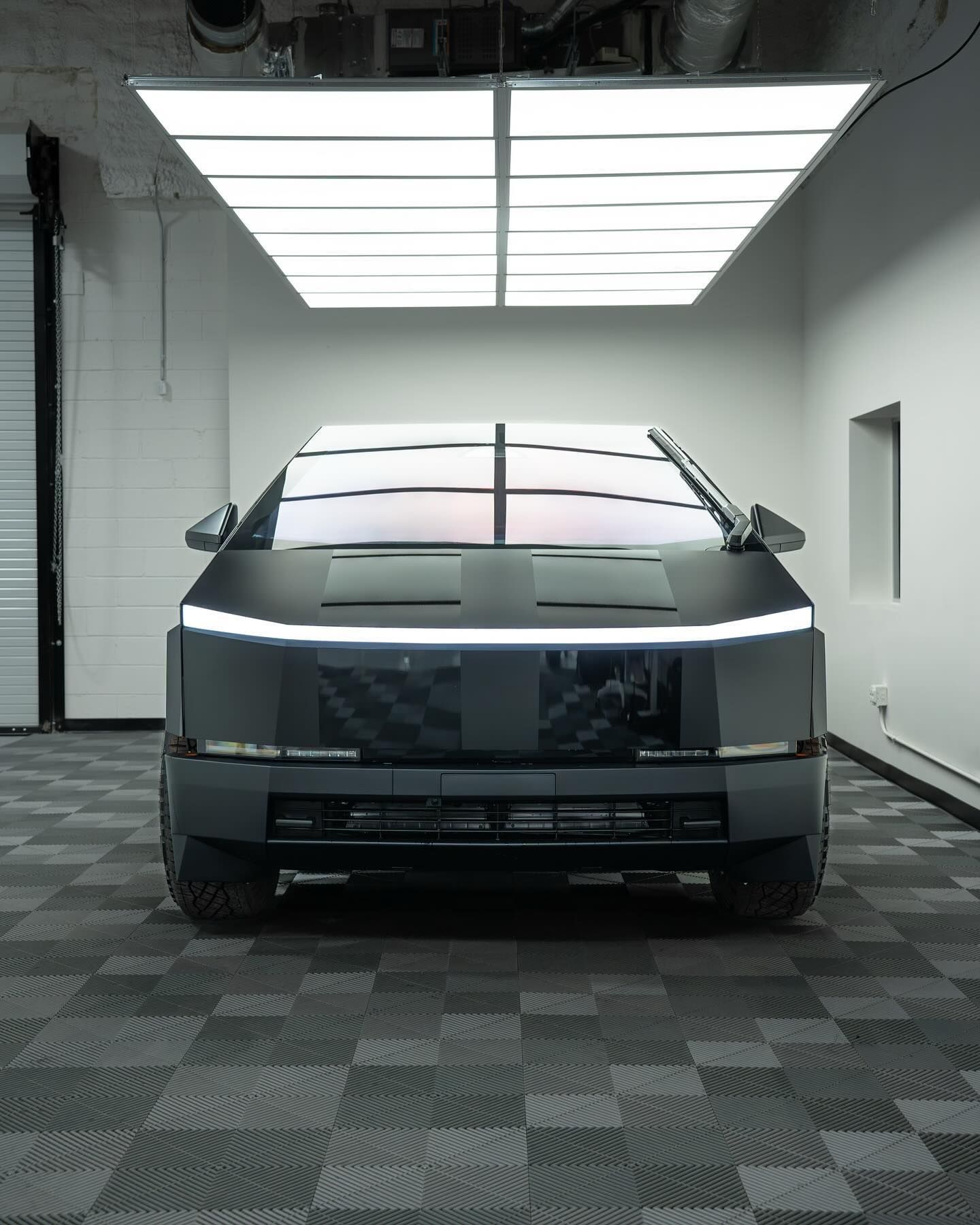 A matte black, angular Cybertruck parked in a bright, modern studio with a checkered floor and overhead panel lighting.