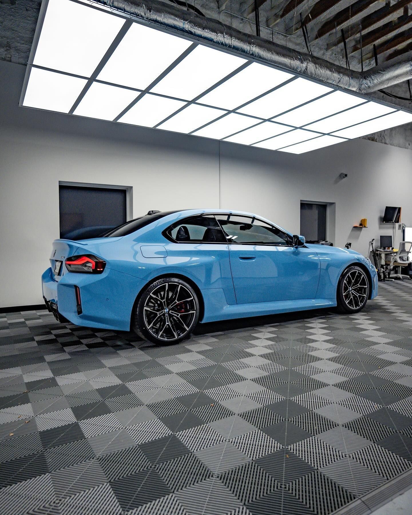 A blue BMW M2 sports car parked inside a brightly lit, modern garage with a checkered floor.