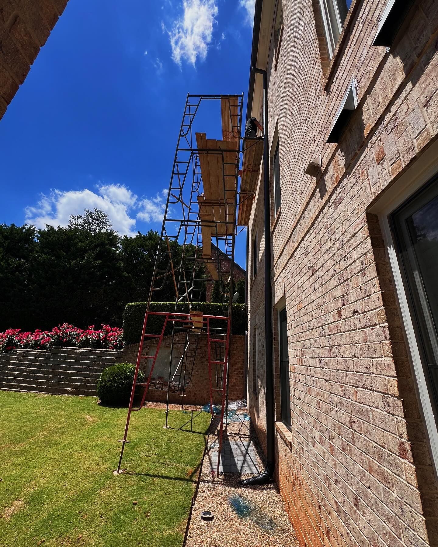 A tall, red metal scaffolding tower stands against the brick wall of a house on a sunny day in a grassy backyard.