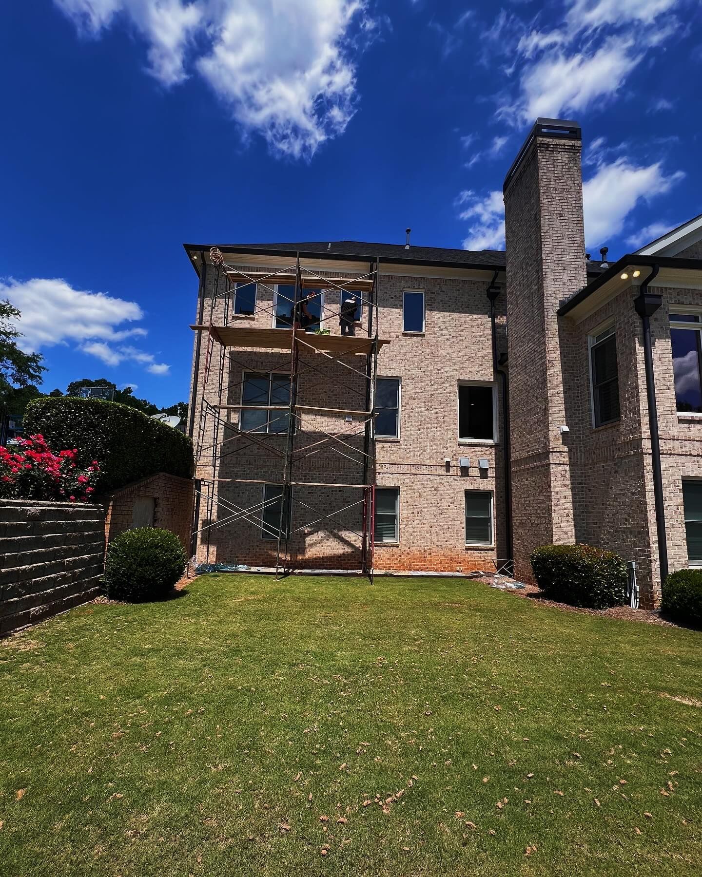 A multi-story brick house with a tall stone chimney and metal scaffolding against the exterior wall in a backyard.