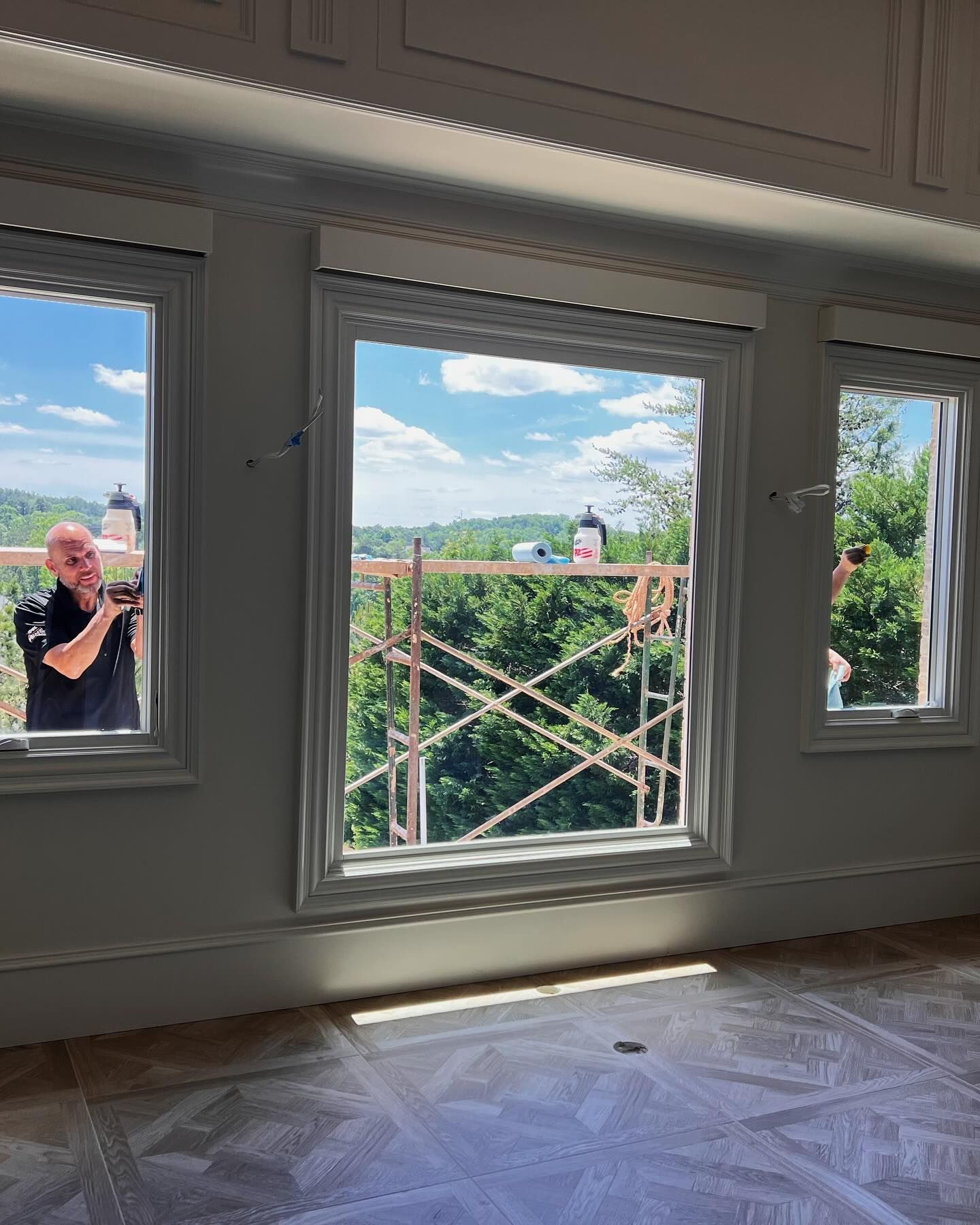 A person leans into an open window to work, overlooking scaffolding against trees, inside a room with patterned flooring.