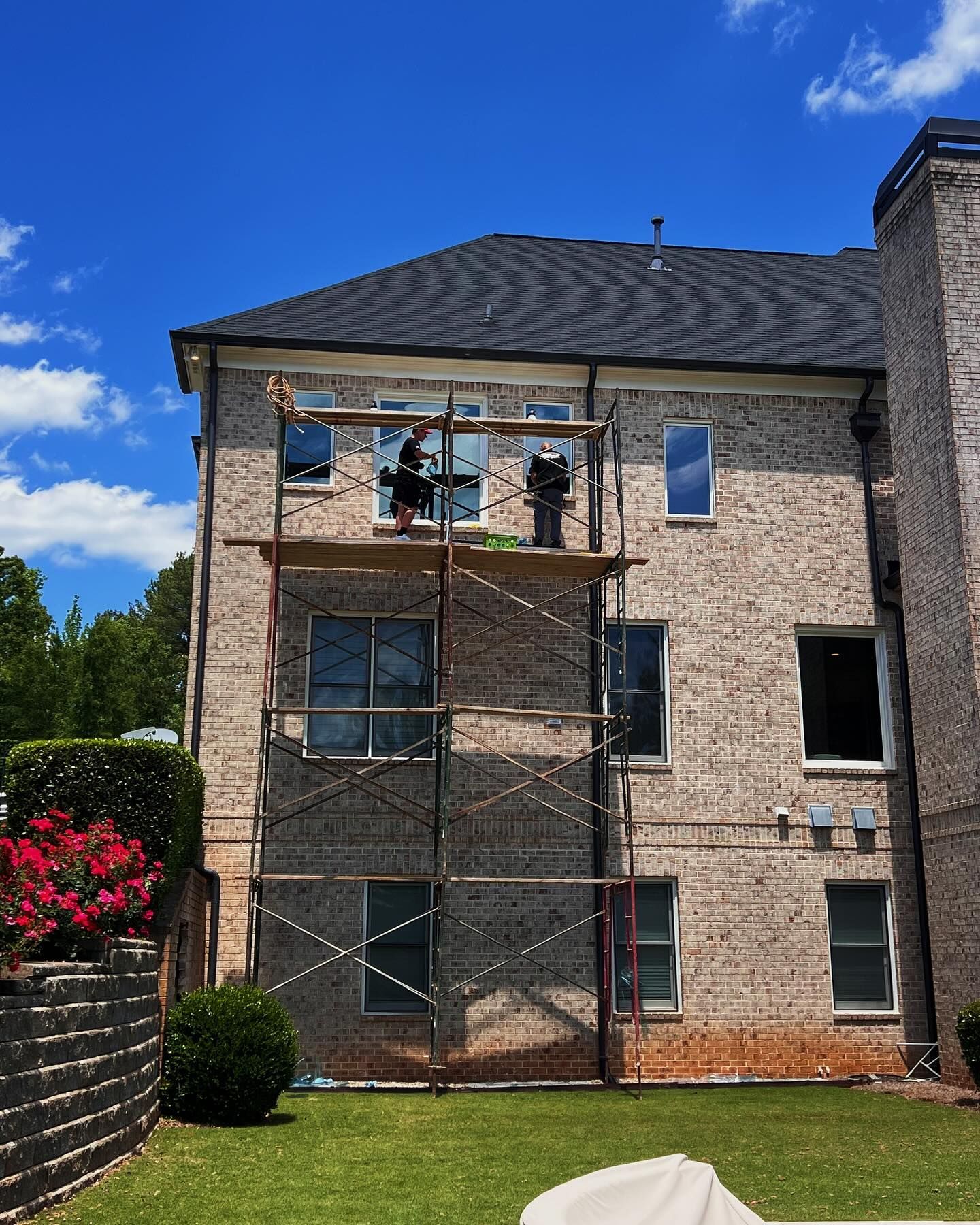 Two workers on scaffolding perform exterior home maintenance on the brick wall of a two-story house under a blue sky.