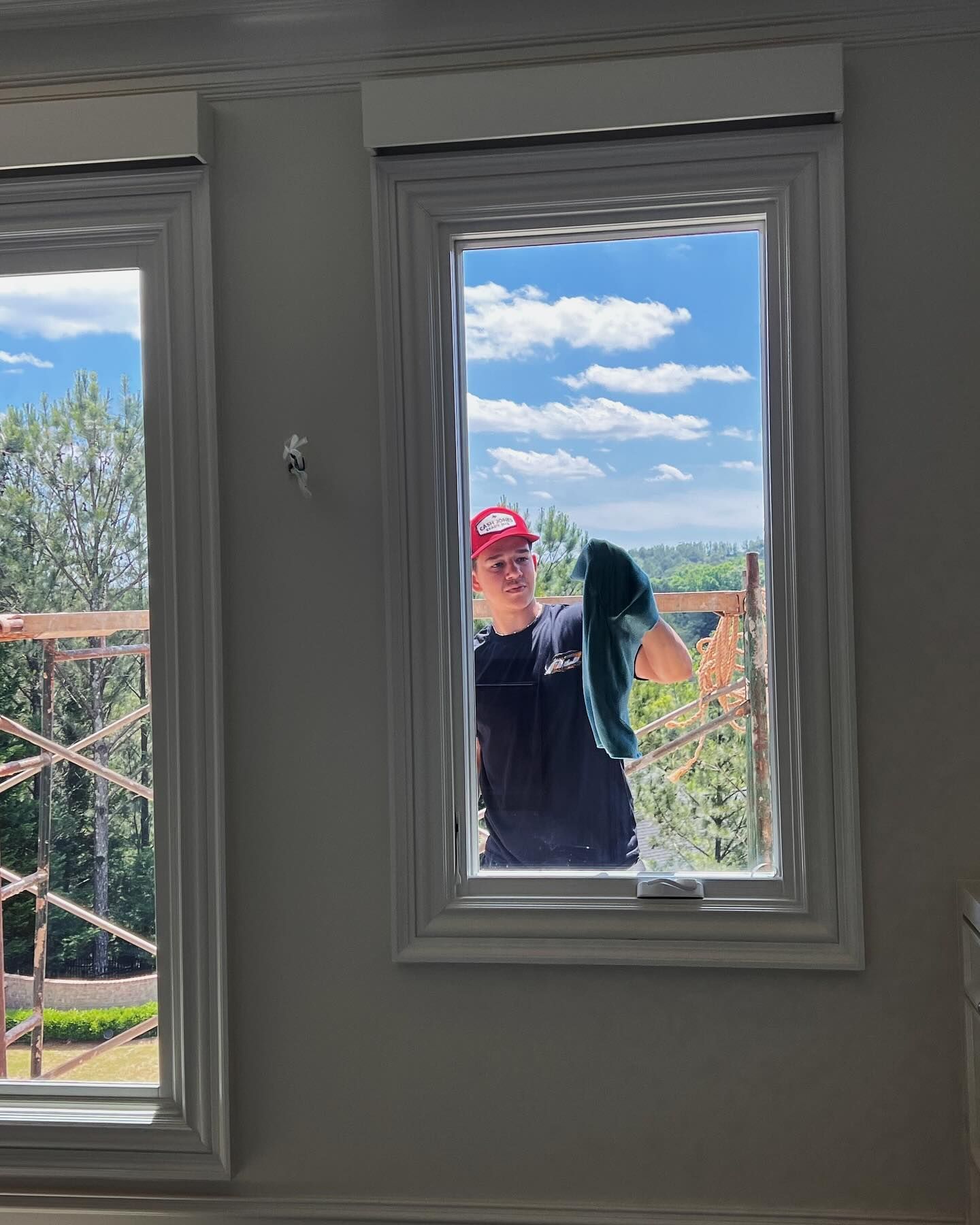A person wearing a red cap and dark shirt cleaning the exterior of a window while standing on scaffolding.