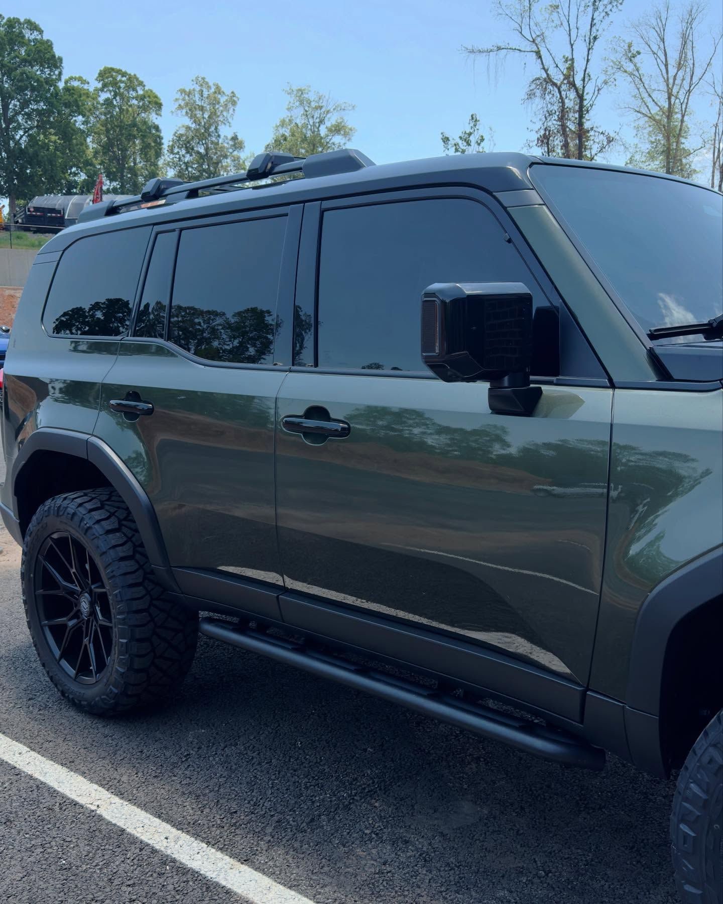 Olive green SUV parked in a lot, featuring tinted windows, black wheels, and off-road tires.