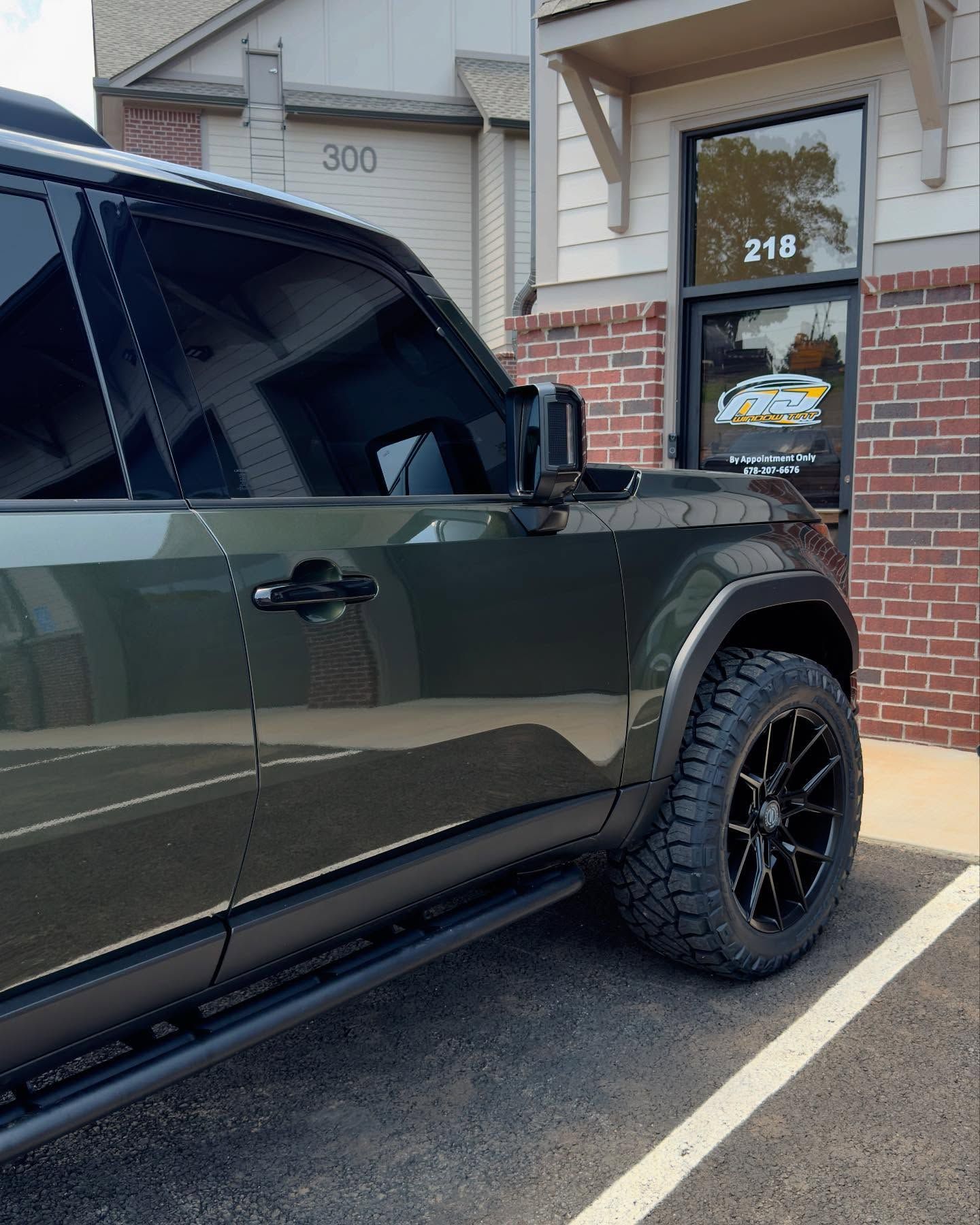 Close-up side view of an olive green SUV with black off-road wheels and tinted windows parked in front of a brick building.