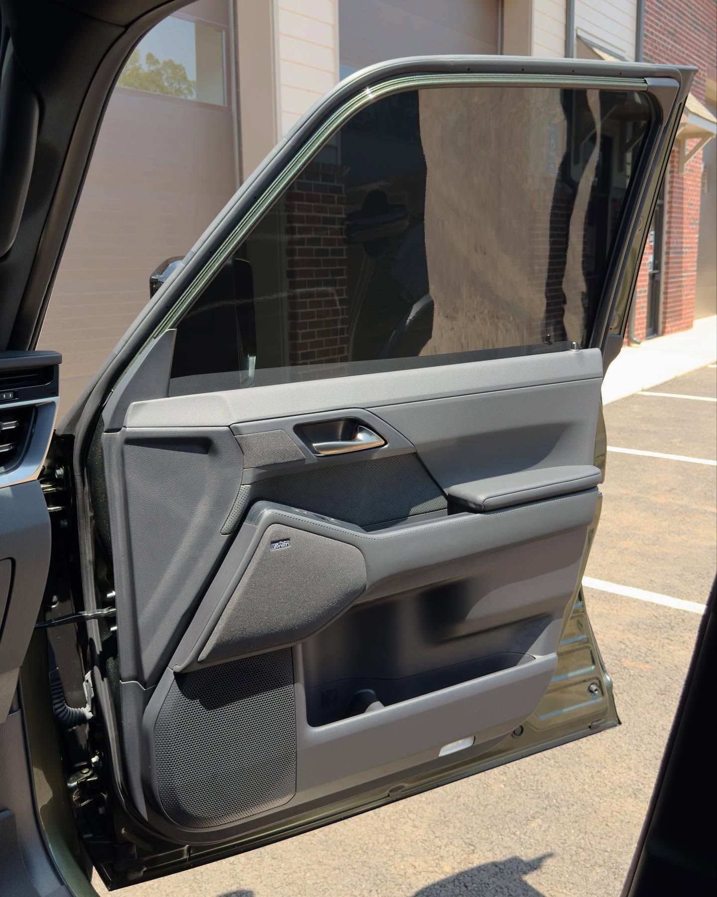 A close-up interior view of a modern vehicle door panel, featuring dark gray trim and a speaker, with a tinted window.
