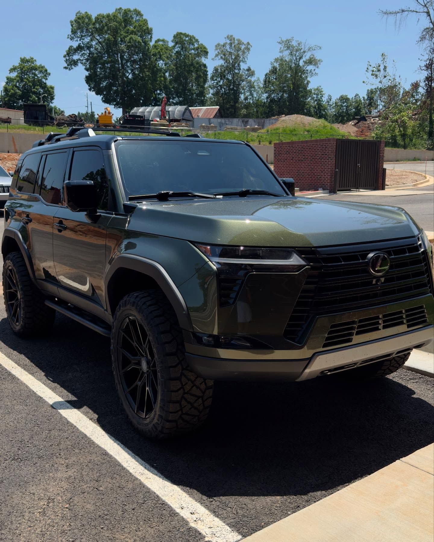 Olive green Lexus GX SUV with black off-road wheels parked on an asphalt lot on a sunny day.