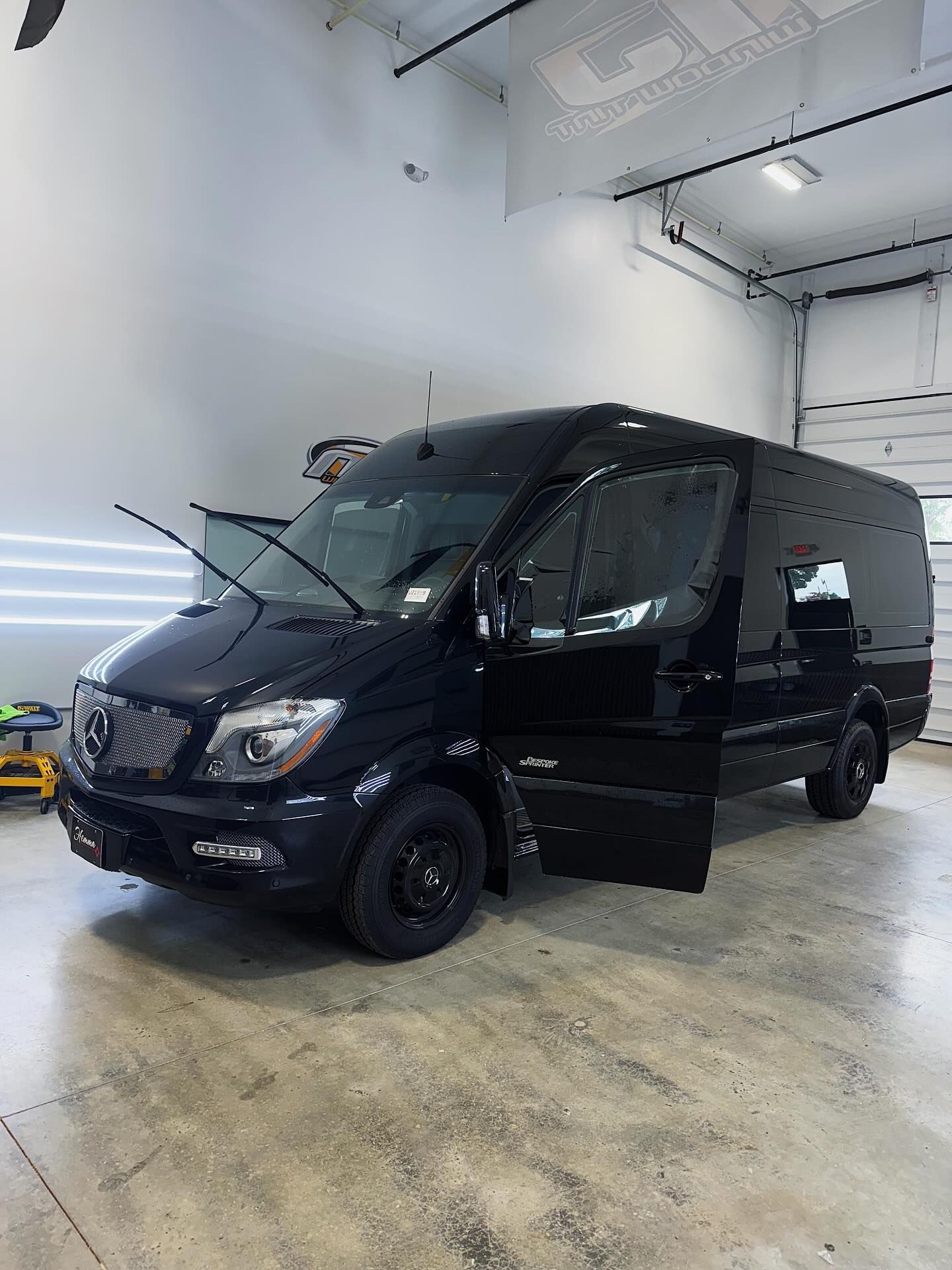 A black Mercedes-Benz Sprinter cargo van with open front doors parked inside a bright, industrial garage.