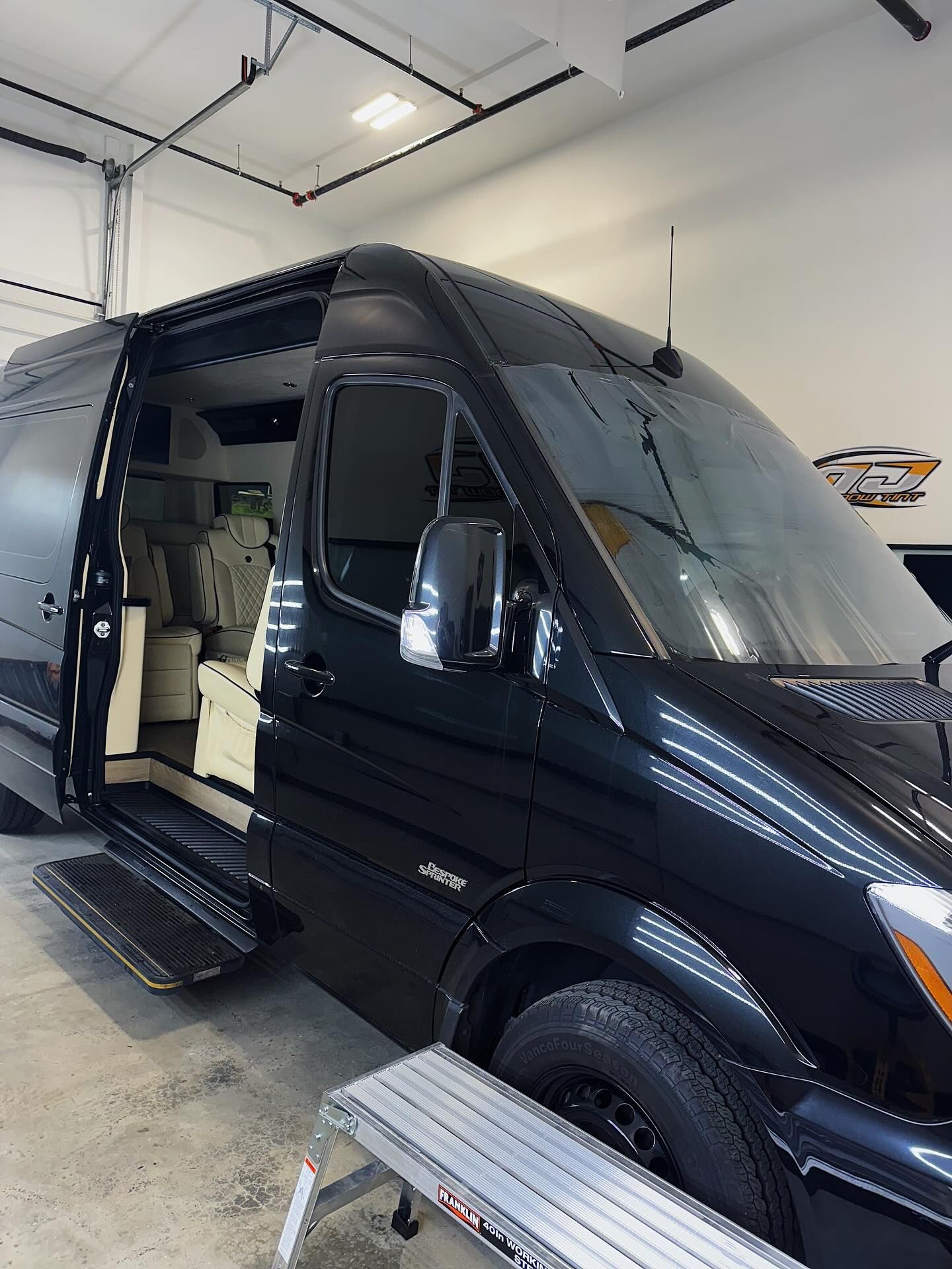 A black Mercedes-Benz Sprinter van parked indoors with its side passenger door open, revealing a luxurious interior.