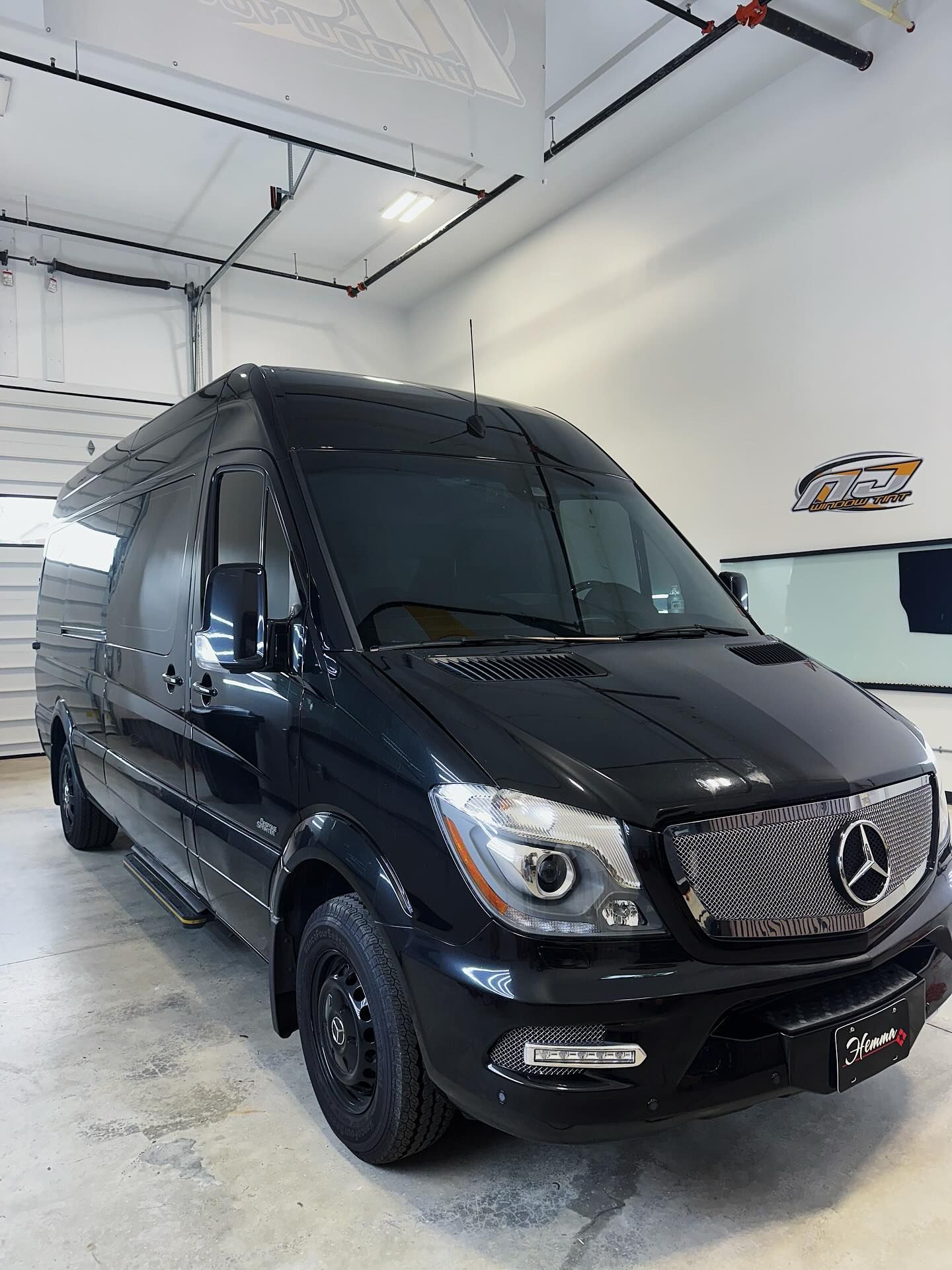 A black Mercedes-Benz Sprinter van parked inside a bright, clean garage with white walls and industrial lighting.