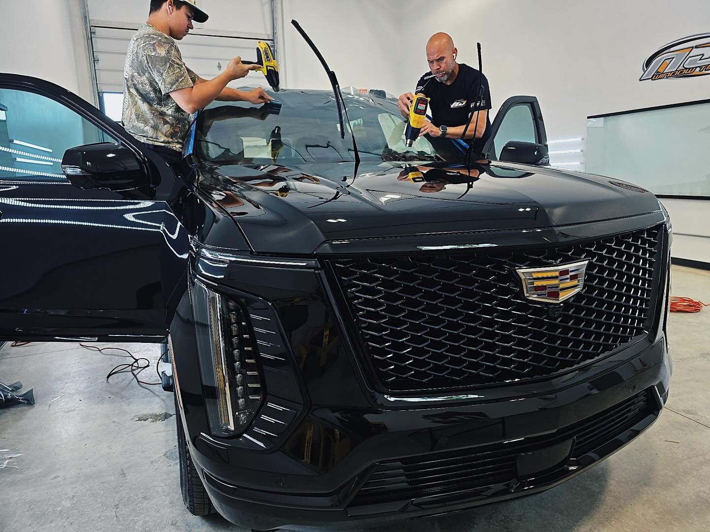 Two technicians in a garage use handheld tools to install protective film on the windshield of a black Cadillac SUV.