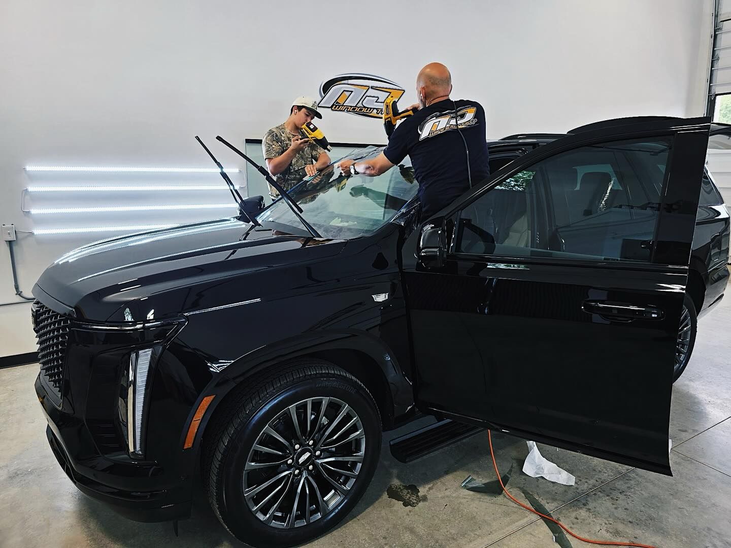 Two technicians work on installing a new windshield on a black Cadillac SUV inside a professional service garage.