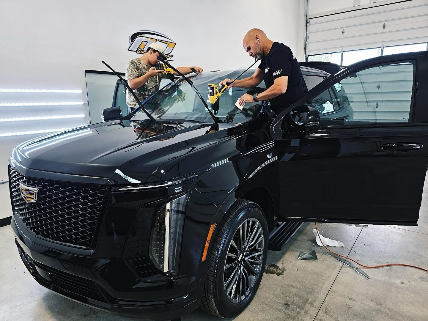 Two technicians work in a professional shop to apply window tint to the windshield of a black Cadillac SUV.