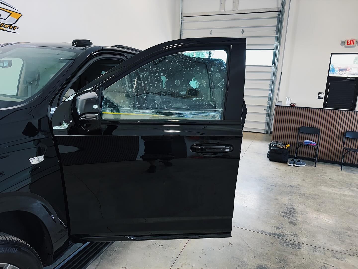 A black truck door is open inside a garage, with clear protective film being applied to the window glass.