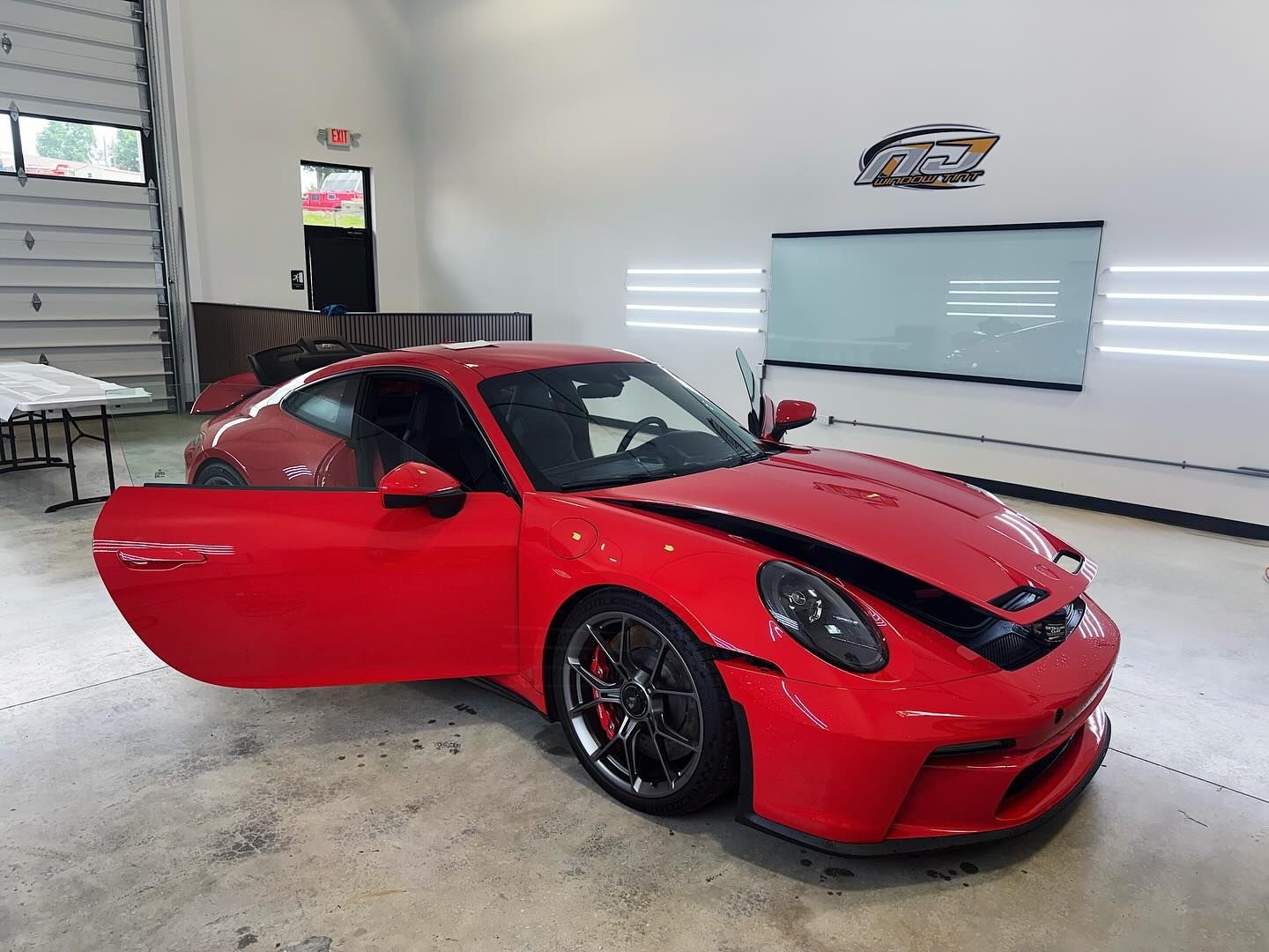 A red Porsche 911 GT3 with its driver-side door open, parked inside a bright, clean auto shop.