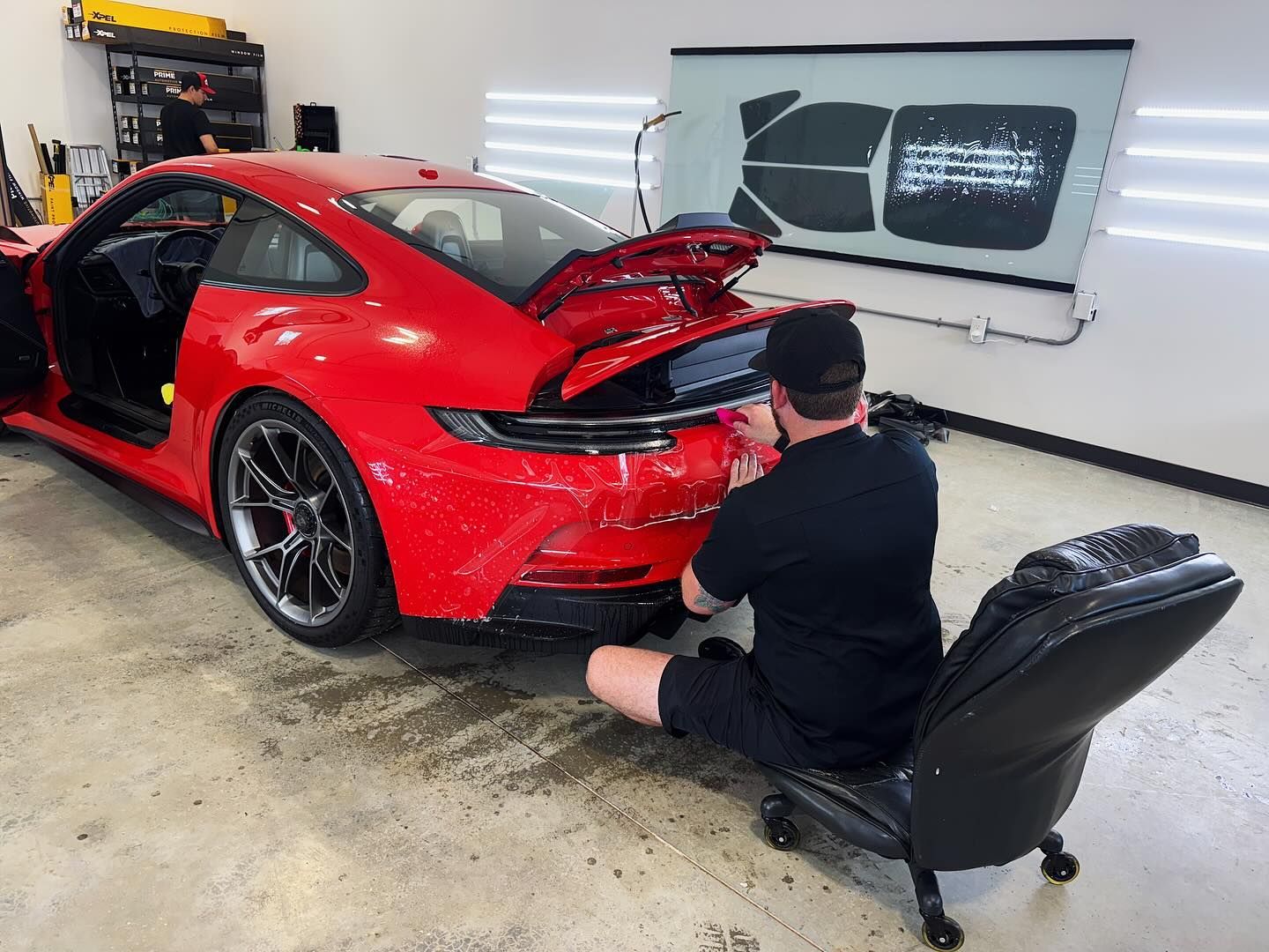 A worker in a shop uses a rolling chair to detail the rear of a bright red Porsche 911 with its engine cover open.