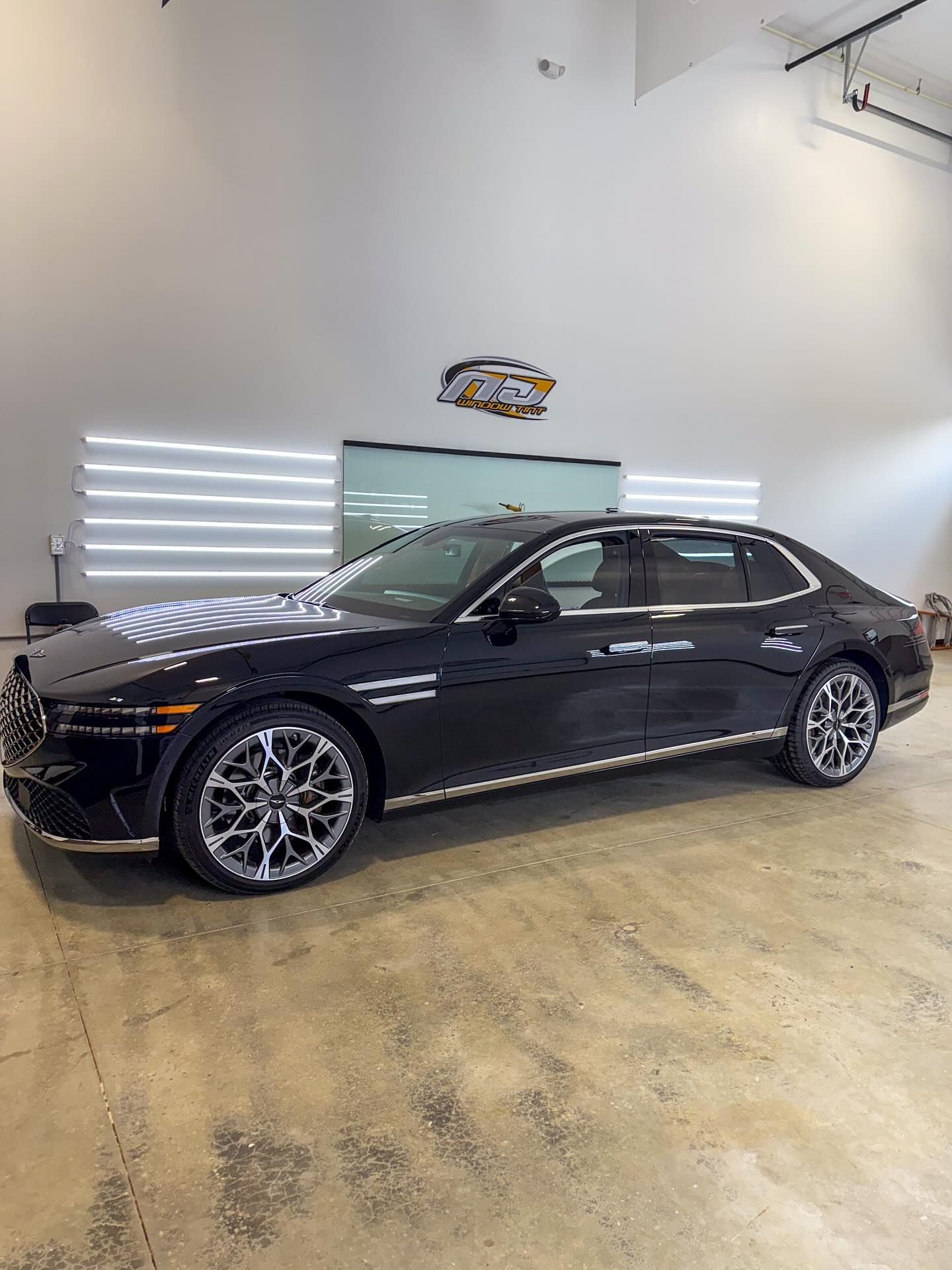 A black Genesis G80 sedan parked in a bright, modern indoor garage with white accent lighting.