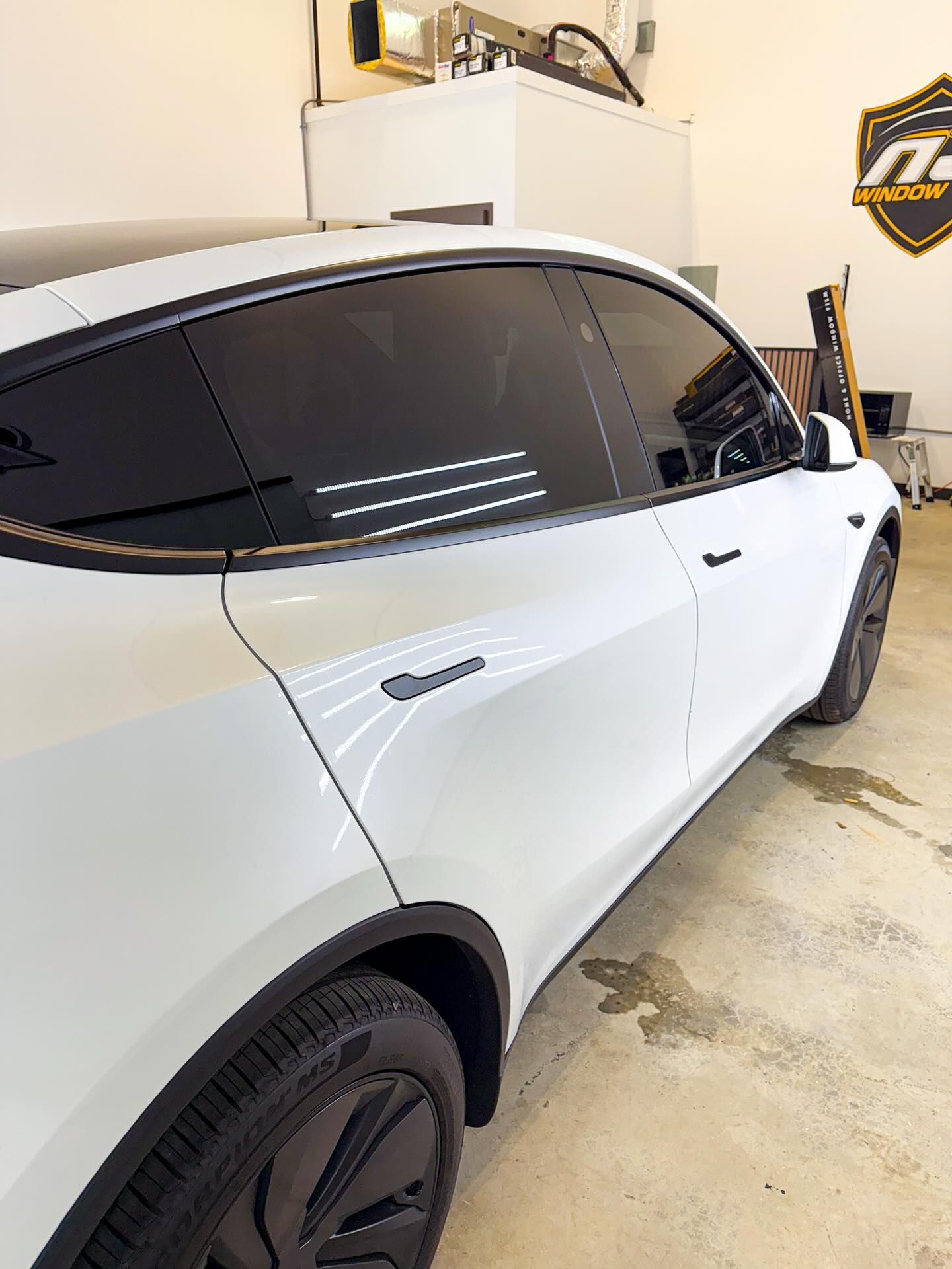 A white Tesla Model Y in a garage, featuring dark tinted windows and black wheels, viewed from the rear passenger side.