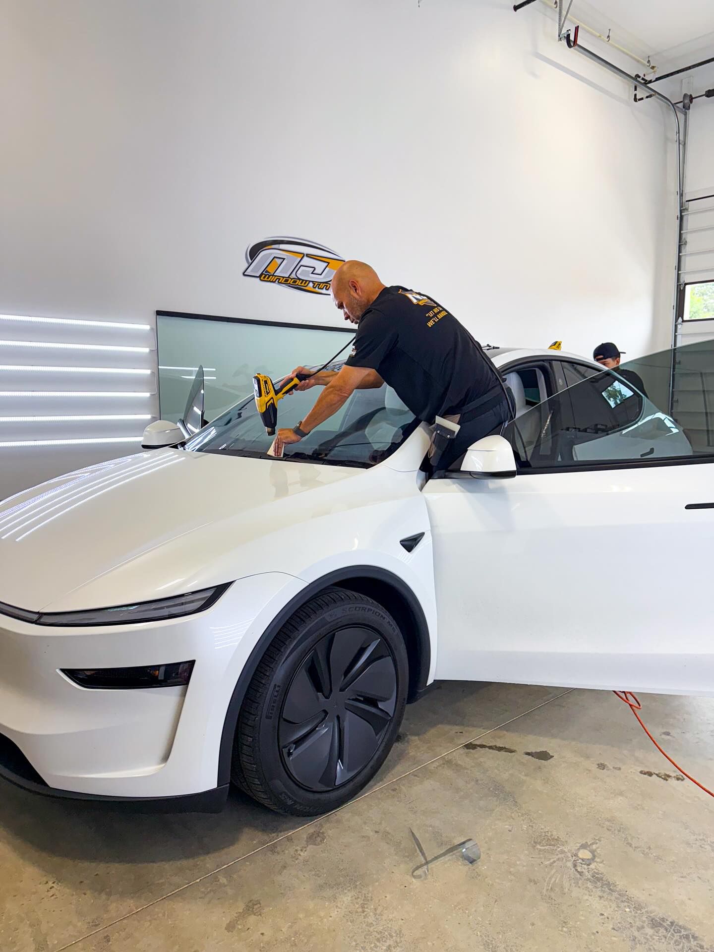 A technician uses a heat gun to apply window tint to the windshield of a white Tesla in an indoor workshop.