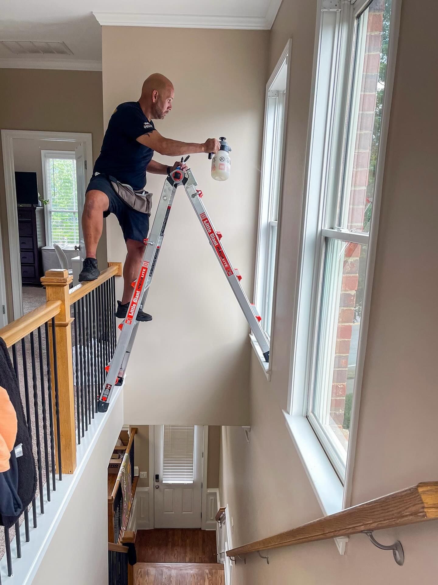 A person balances a folding ladder between a staircase railing and a window ledge to spray-paint the wall.