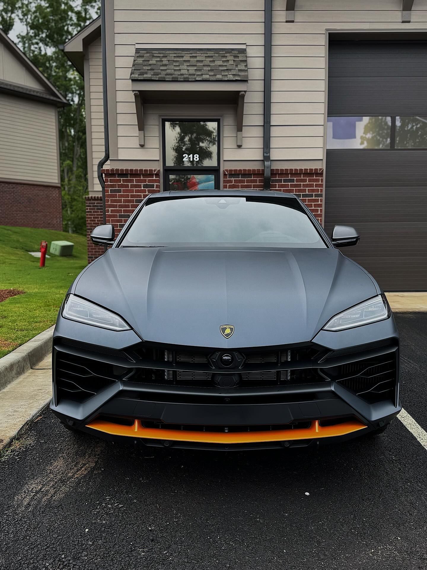 A matte gray Lamborghini Urus with a bright orange front splitter, parked on a paved driveway in front of a house.