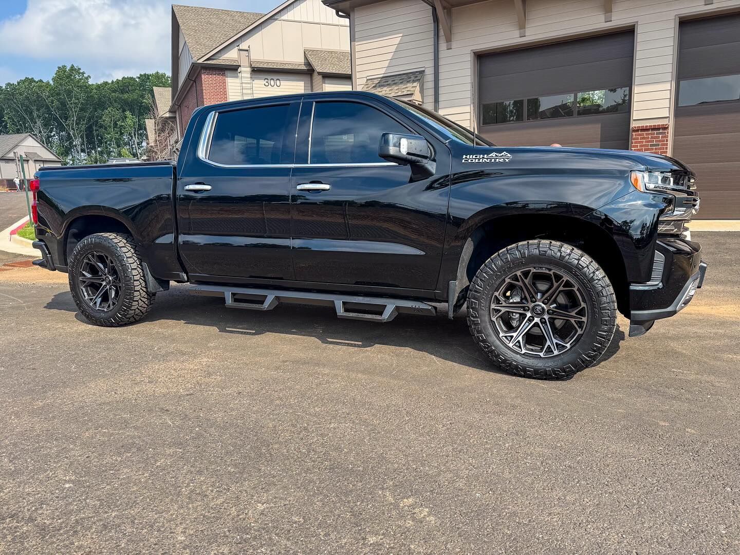 A black Chevrolet Silverado pickup truck with custom wheels parked on an asphalt driveway in front of a house.