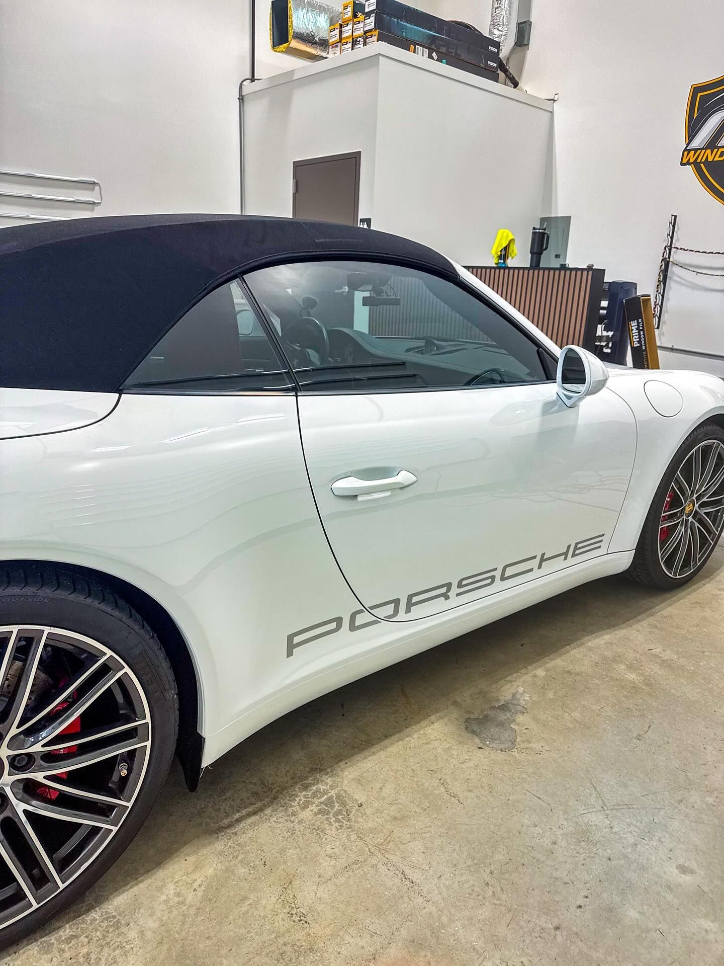 A white Porsche convertible with a black soft top and black wheels, parked inside a garage or shop.
