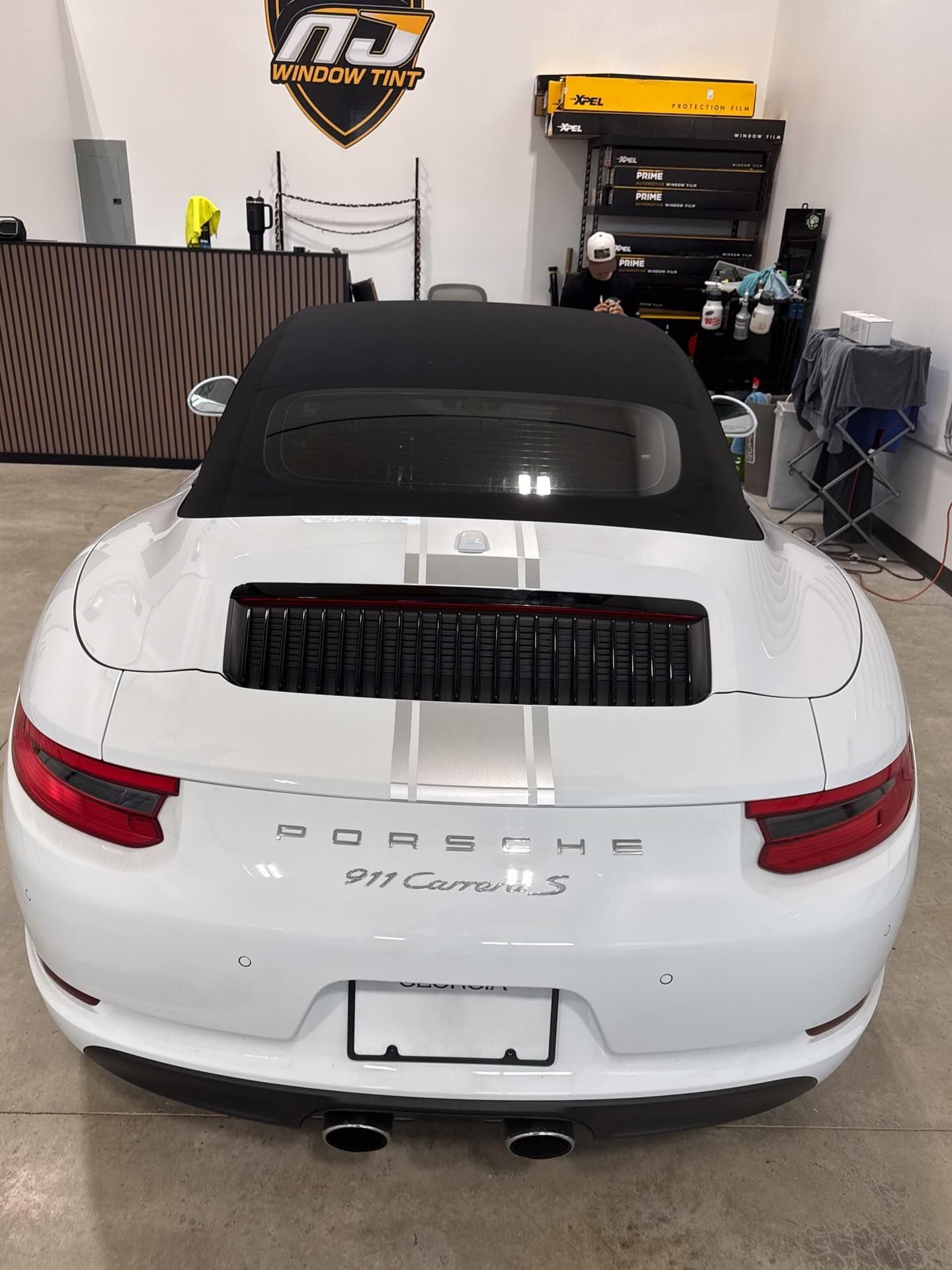 White Porsche 911 Carrera S convertible with black soft top, centered in a garage with a logo on the wall.