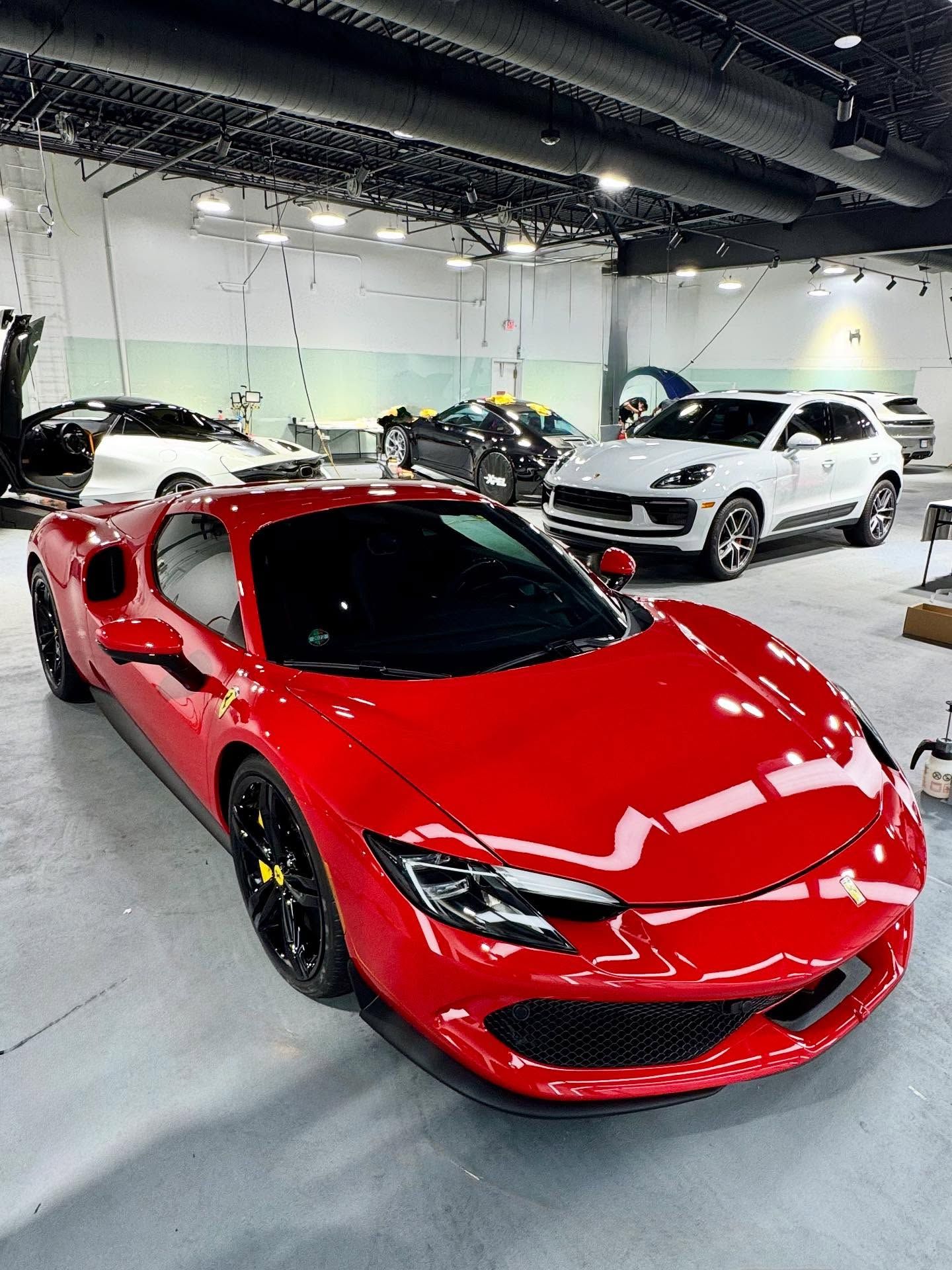 A bright red Ferrari sports car parked in the foreground of a large, well-lit indoor vehicle showroom.