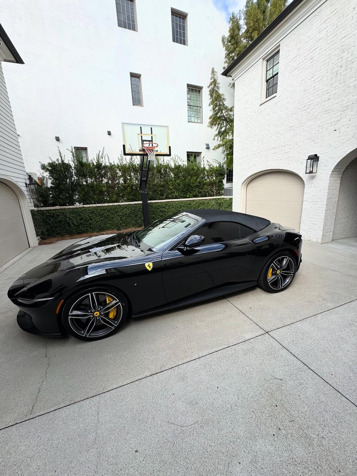A black Ferrari convertible parked in a white stone driveway next to a residential building with a basketball hoop.