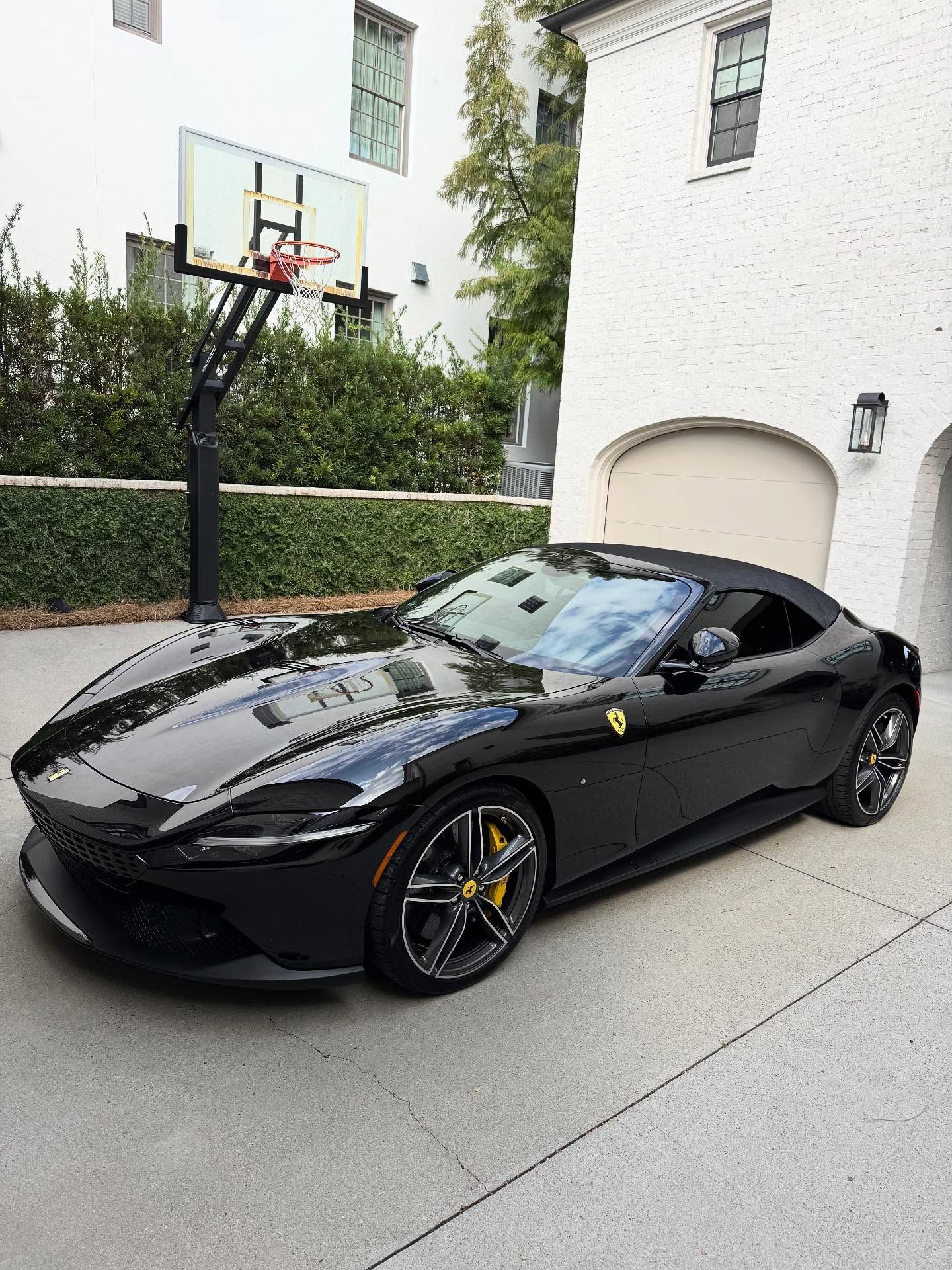 A black Ferrari convertible parked on a driveway in front of a white house with a basketball hoop.