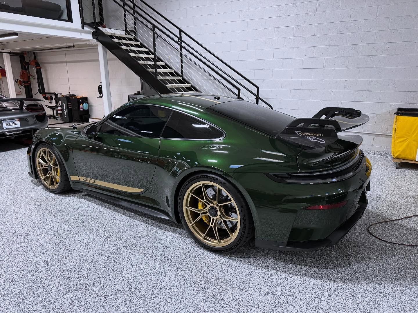 A dark green Porsche 911 GT3 with gold wheels parked in a modern garage with a staircase and light-colored flooring.