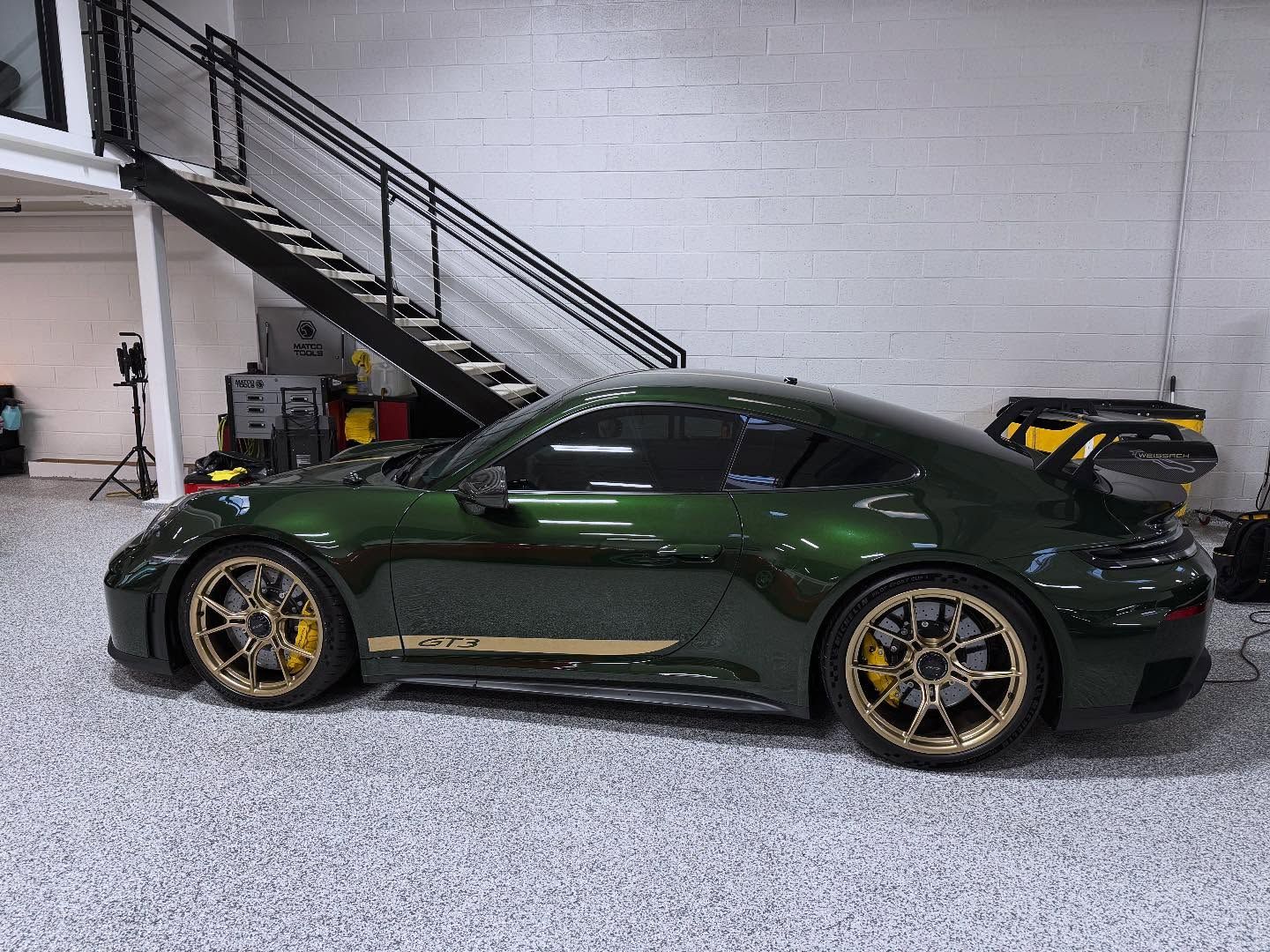 Dark green Porsche 911 GT3 with gold wheels and decals, parked inside a garage with a staircase in the background.