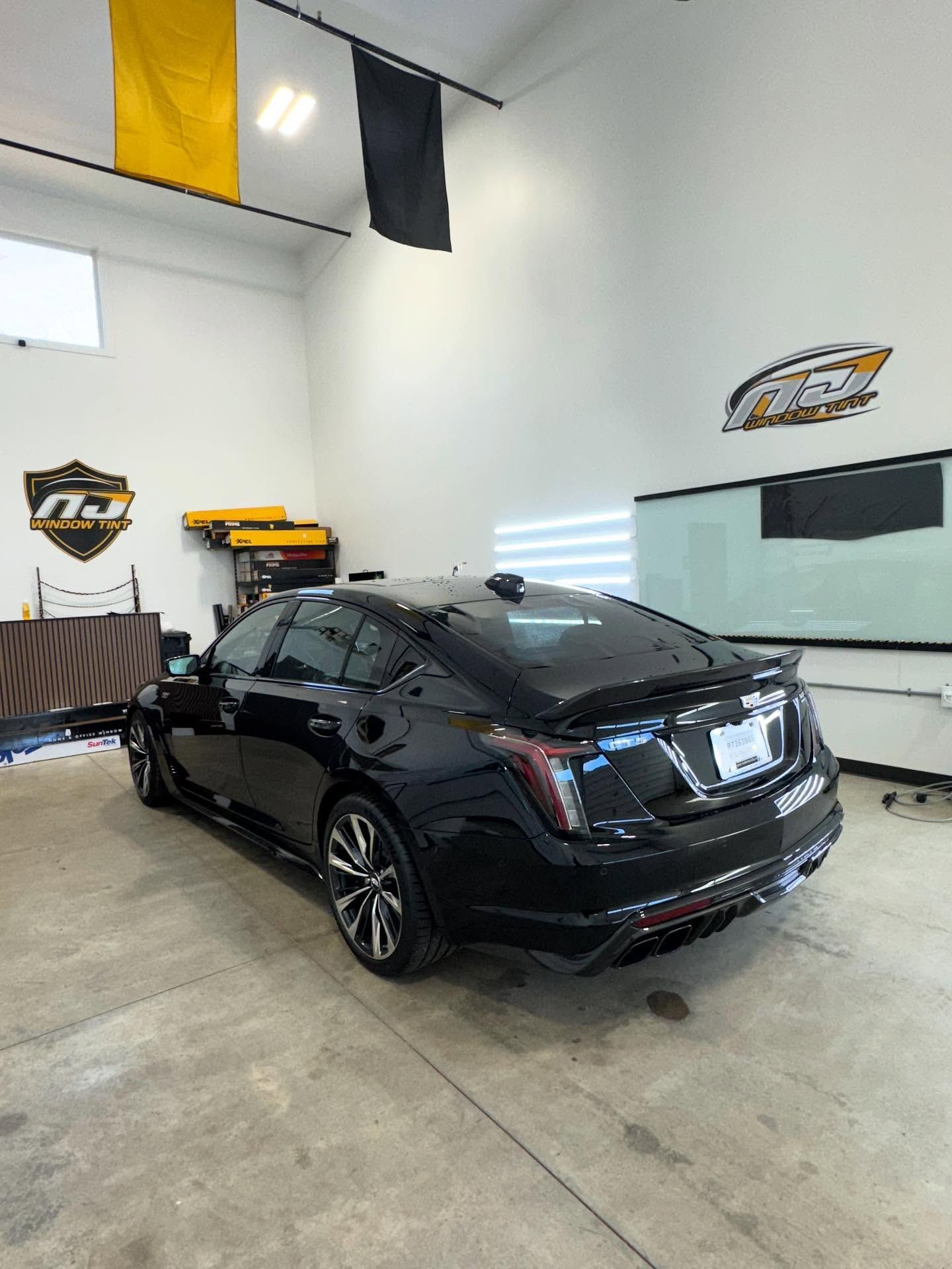 A black Cadillac CT5-V parked inside a professional automotive garage with branded banners and signage on the white walls.