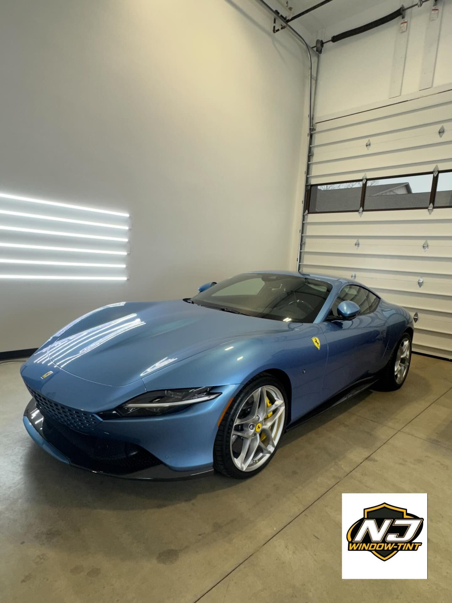 A light blue Ferrari Roma coupe parked in a bright, white garage with a small company logo in the corner.